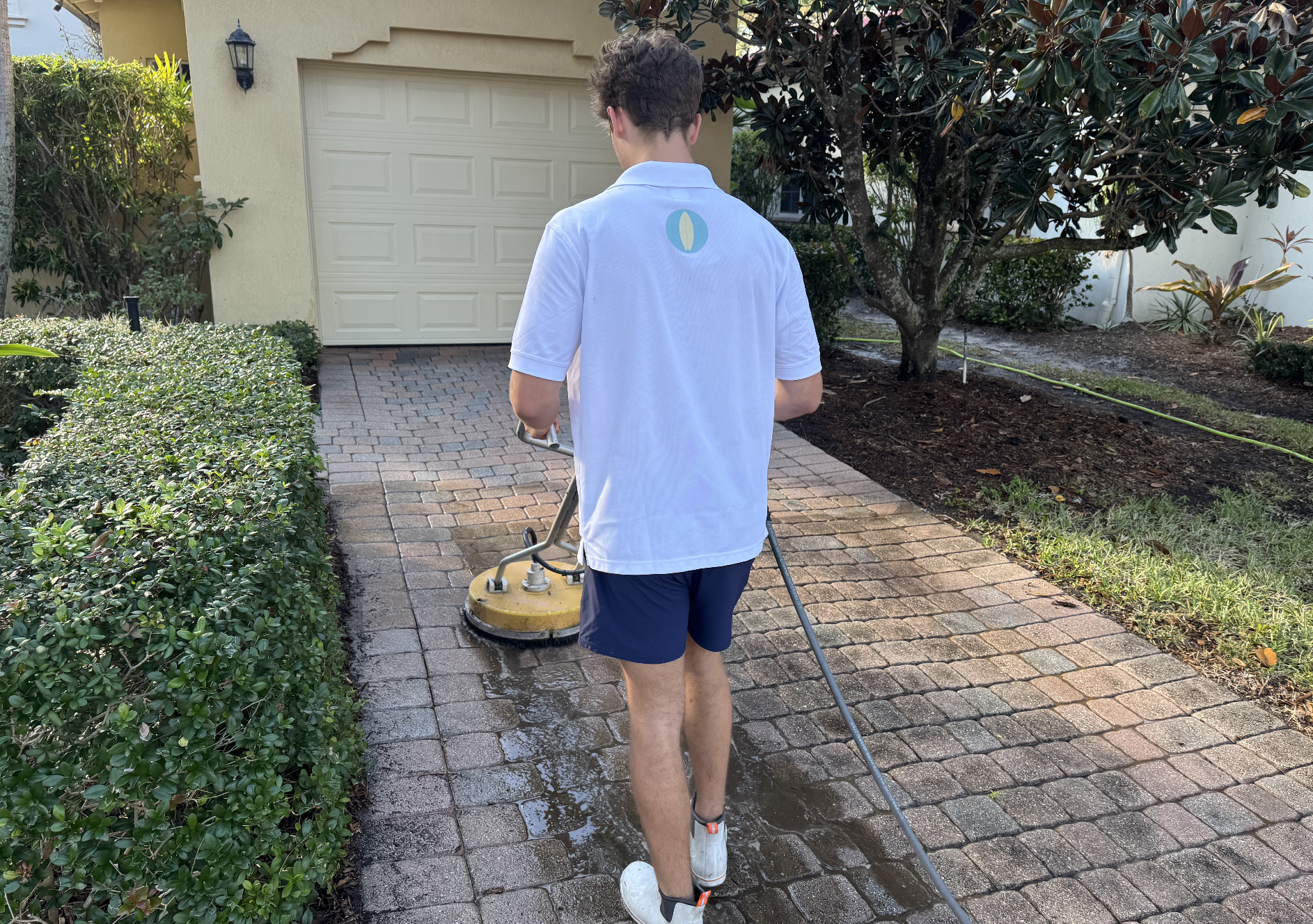 A man wearing a white polo shirt and navy shorts is pressure washing a brick walkway outside a house. The house has a beige garage door, and there are bushes and a tree nearby.