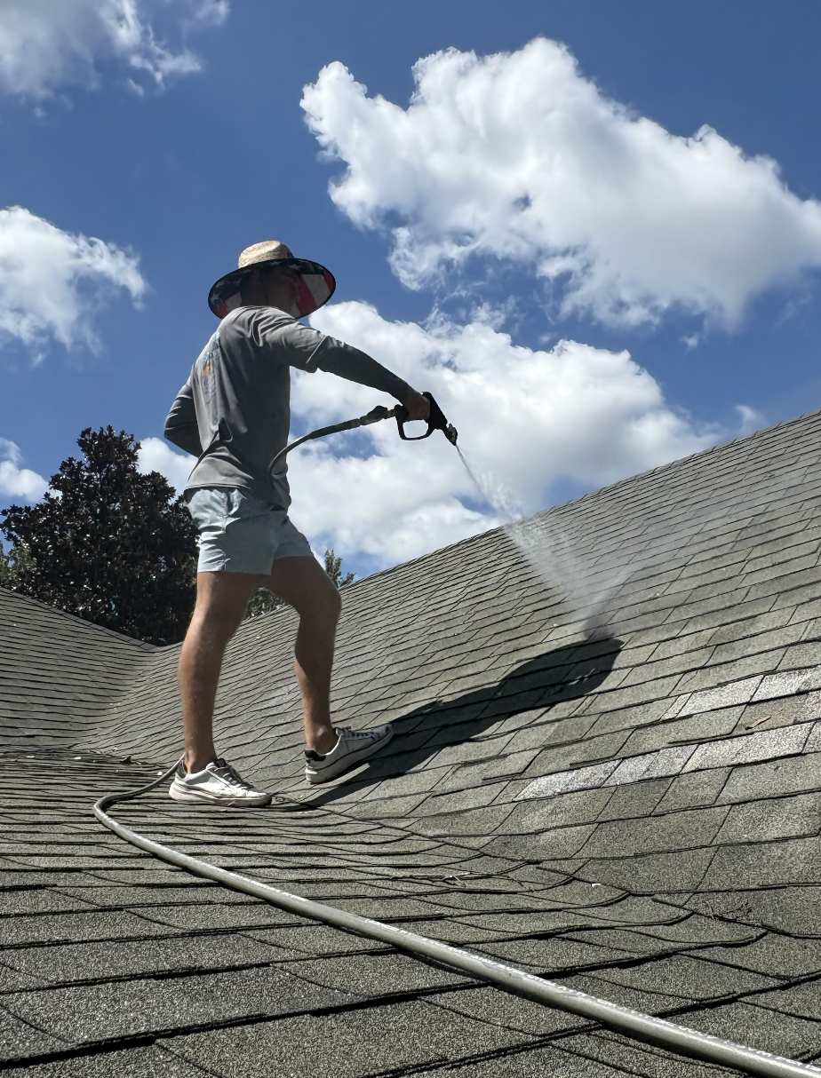 A person wearing a wide-brimmed hat, long-sleeved shirt, and shorts is pressure washing a shingled roof under a partly cloudy sky.