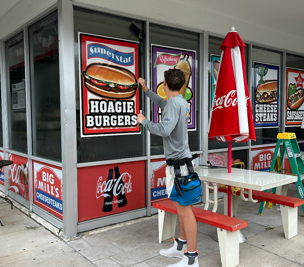 A person in a gray jacket and shorts placing a sign that reads 'Hoagie Burgers' in front of a fast food restaurant window with promotional posters.
