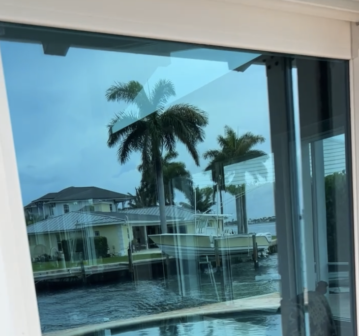 View through a glass window showing palm trees, boats docked at a marina, residential houses, and a partly cloudy sky.