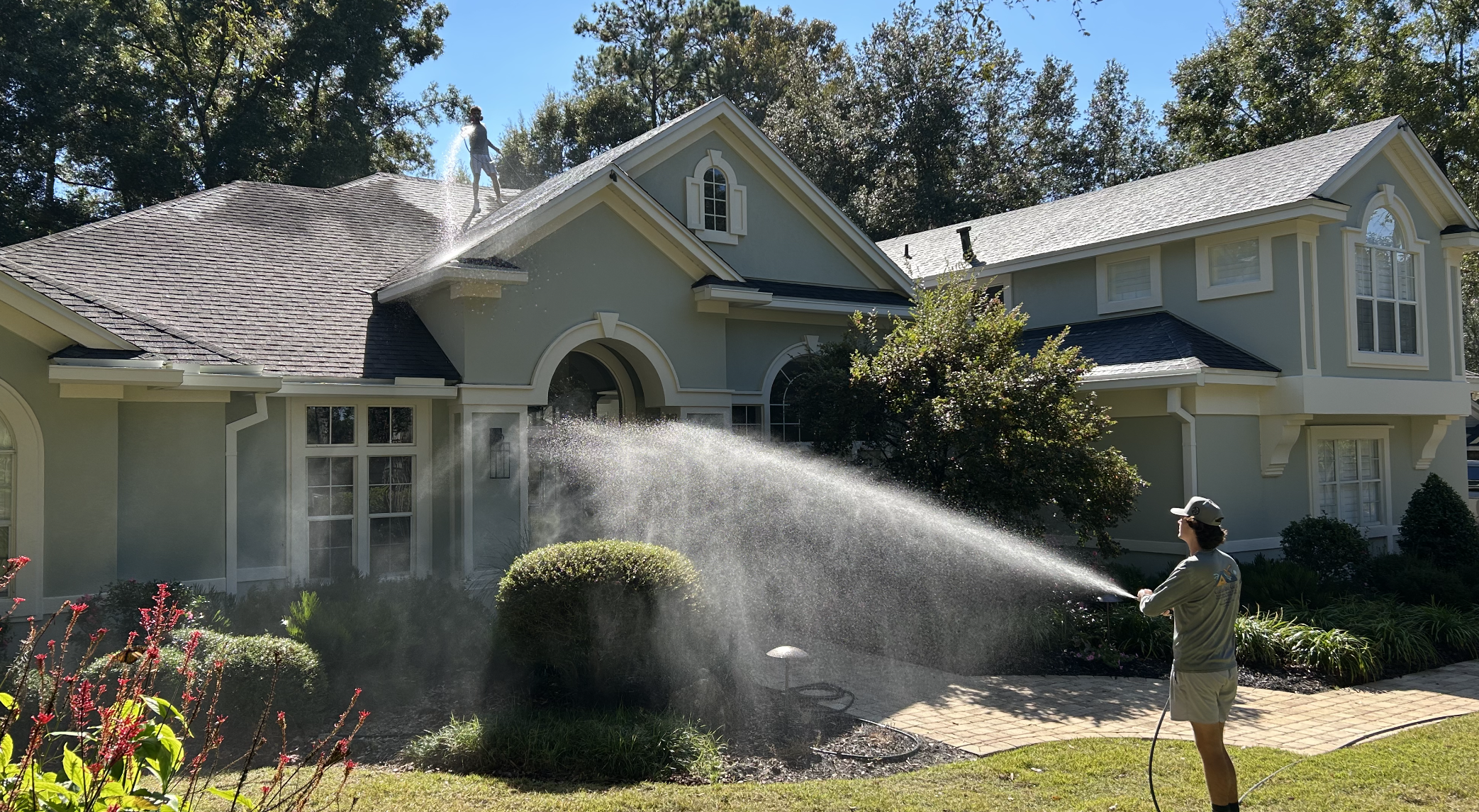 Person watering the lawn with a garden hose in front of a large, light-colored house with multiple windows and gabled roof.