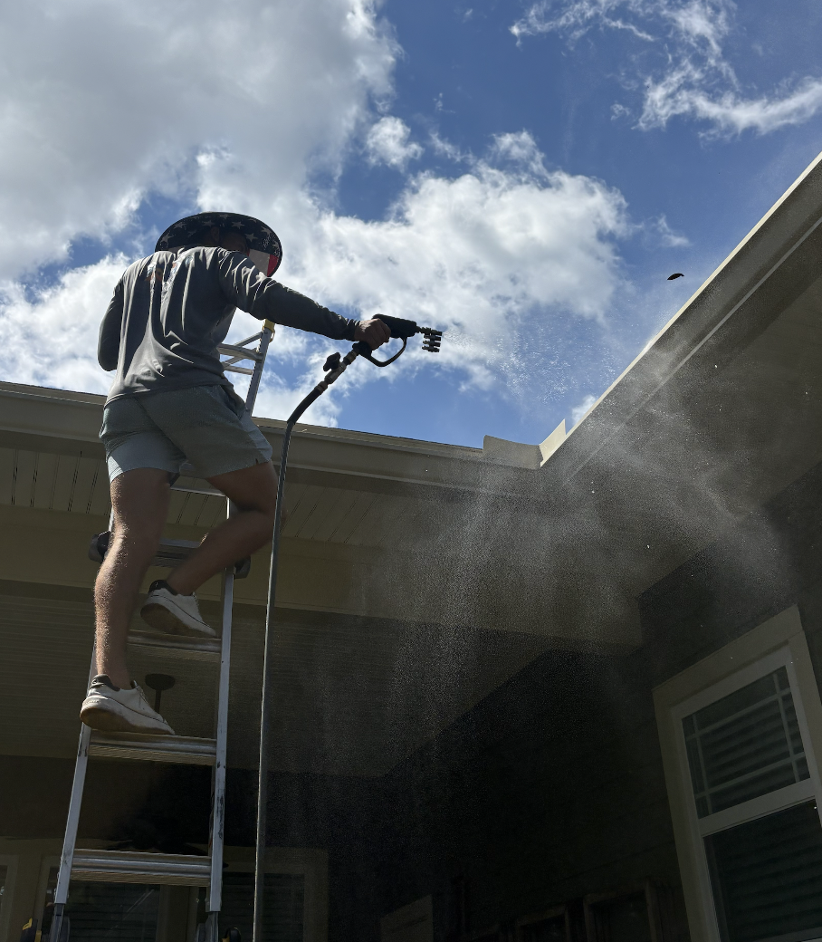 Person standing on a ladder power washing the roof of a house against a blue sky with clouds.