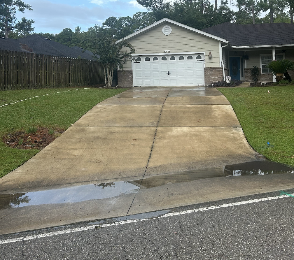 A concrete driveway leading to a white garage door in front of a house with beige siding and gray roof. Wet patches are visible on the driveway, indicating recent rain. The house has a front porch with a blue door, window, and some greenery.