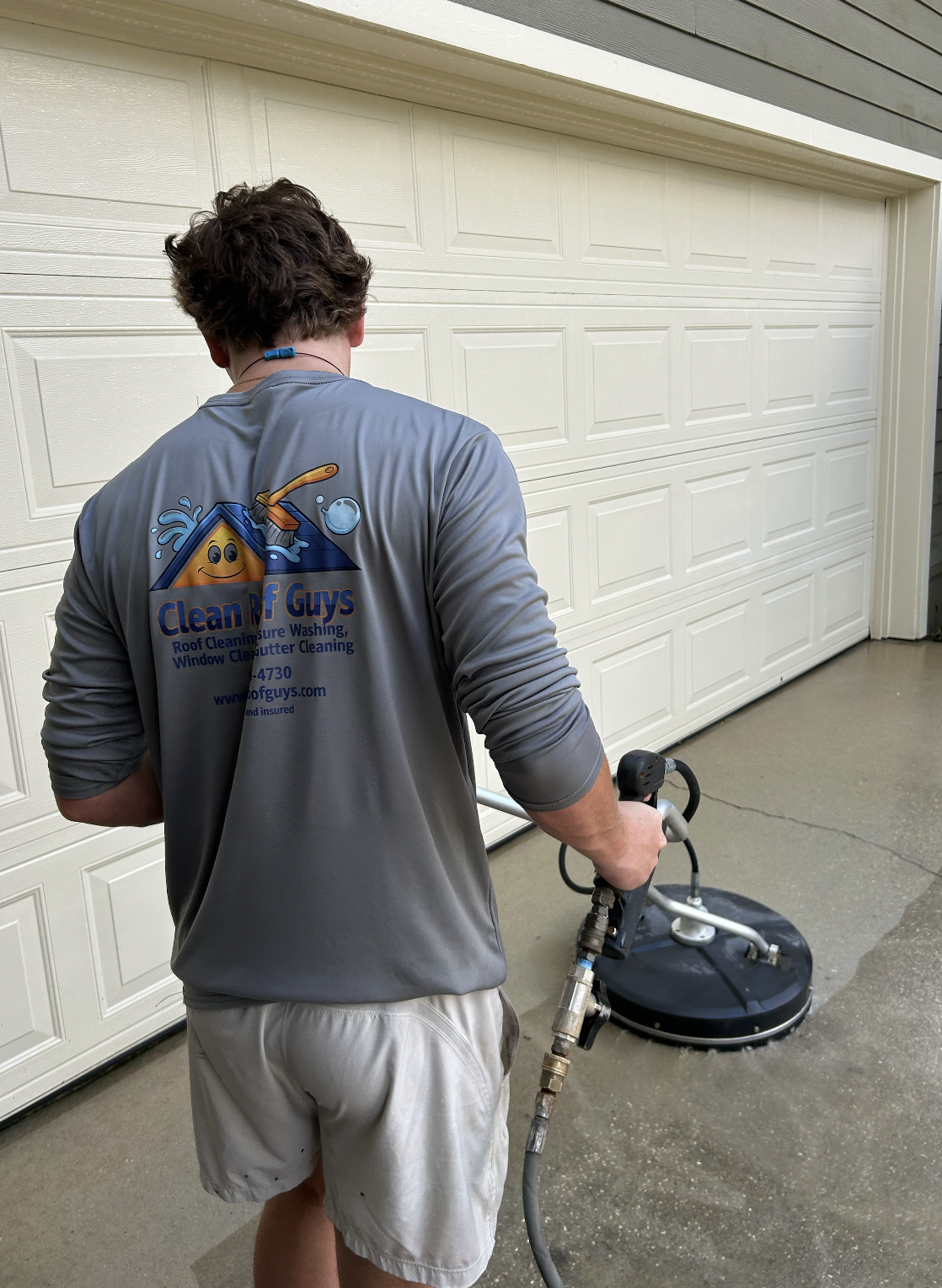 A man in a gray shirt and white shorts pressure washing a garage floor in front of a white garage door.