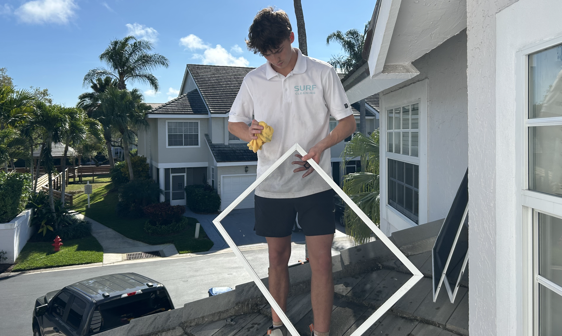 A young man on a roof cleaning a window frame with a yellow cloth, wearing a white shirt with 'SURF CLEANING' printed on it.