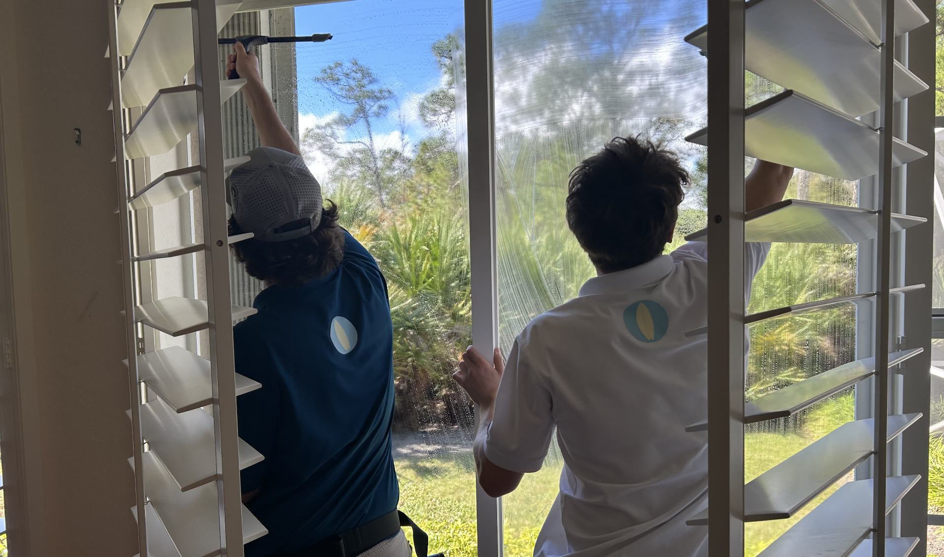 Two individuals cleaning a large window from inside a building, using squeegees to remove water or cleaning solution, with lush greenery outside.