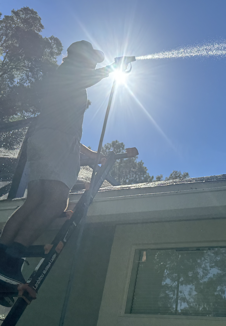 Person cleaning a window outside on a ladder, with the sun shining brightly in the sky.