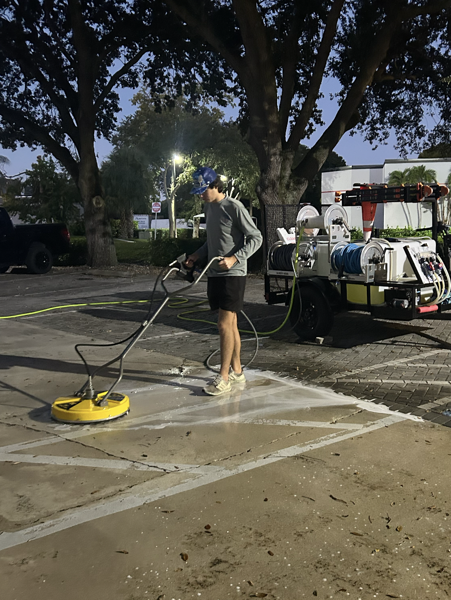 An individual washing a parking lot with a power scrubber machine during twilight, with a wastewater collection trailer nearby and trees in the background.