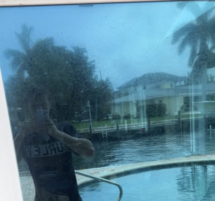Reflected image of a person taking a photo of a backyard pool area through a glass window, with houses, trees, and palm trees in the background.
