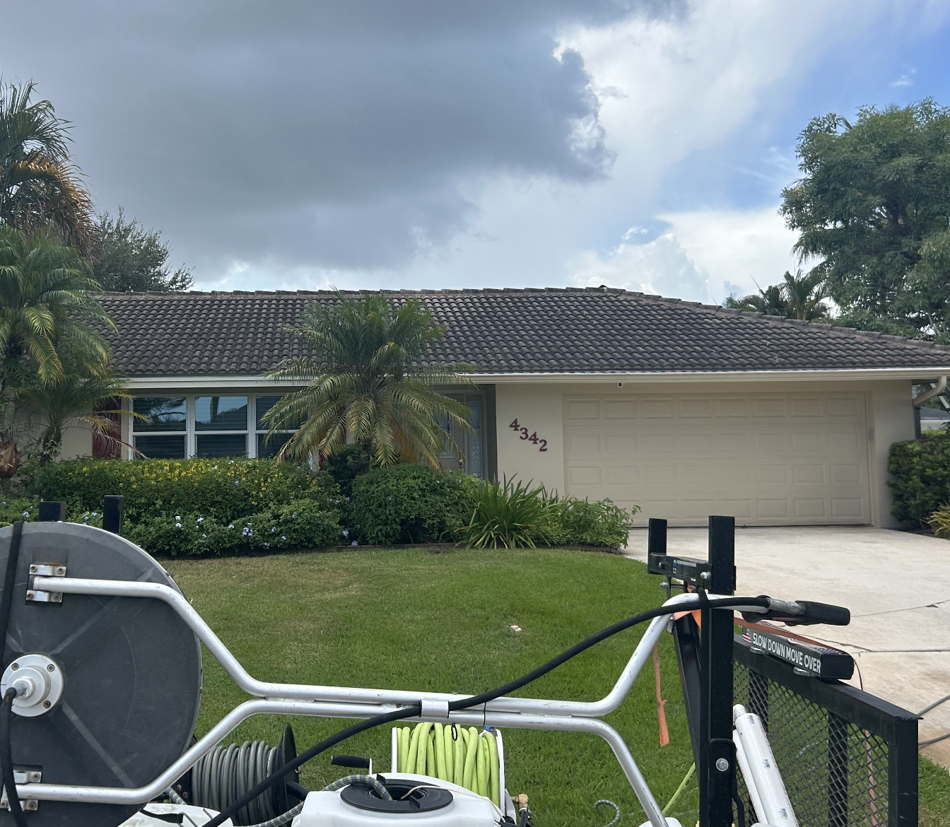 Front view of a house with a brown tile roof, beige garage door, green lawn, and tropical plants, with a cloudy sky overhead. In the foreground, there is a pressure washer and hose on a trailer.