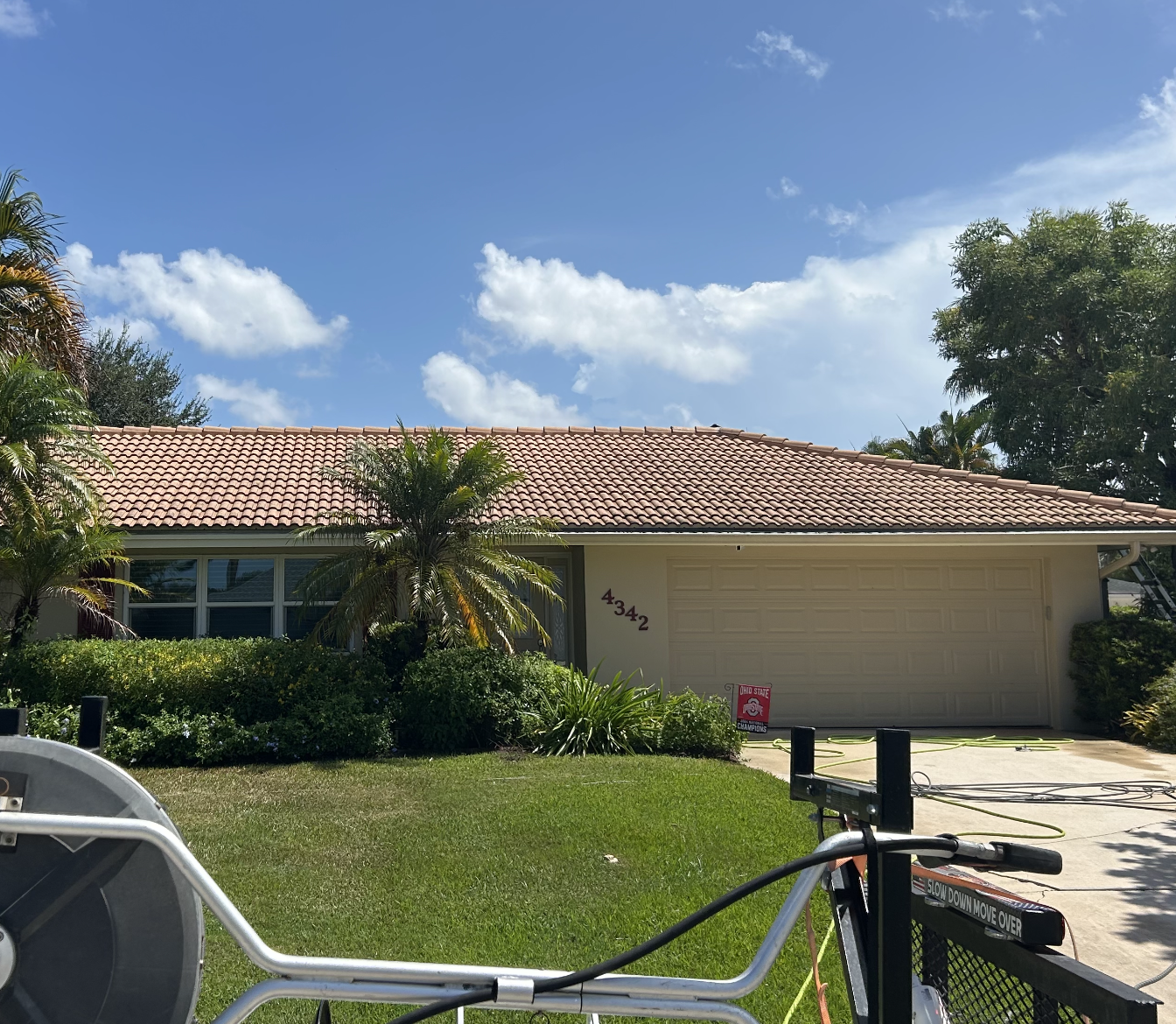 A single-story house with a red-tiled roof, white exterior walls, and a two-car garage. There are palm trees and bushes in the front yard, with a clear blue sky and some clouds above.