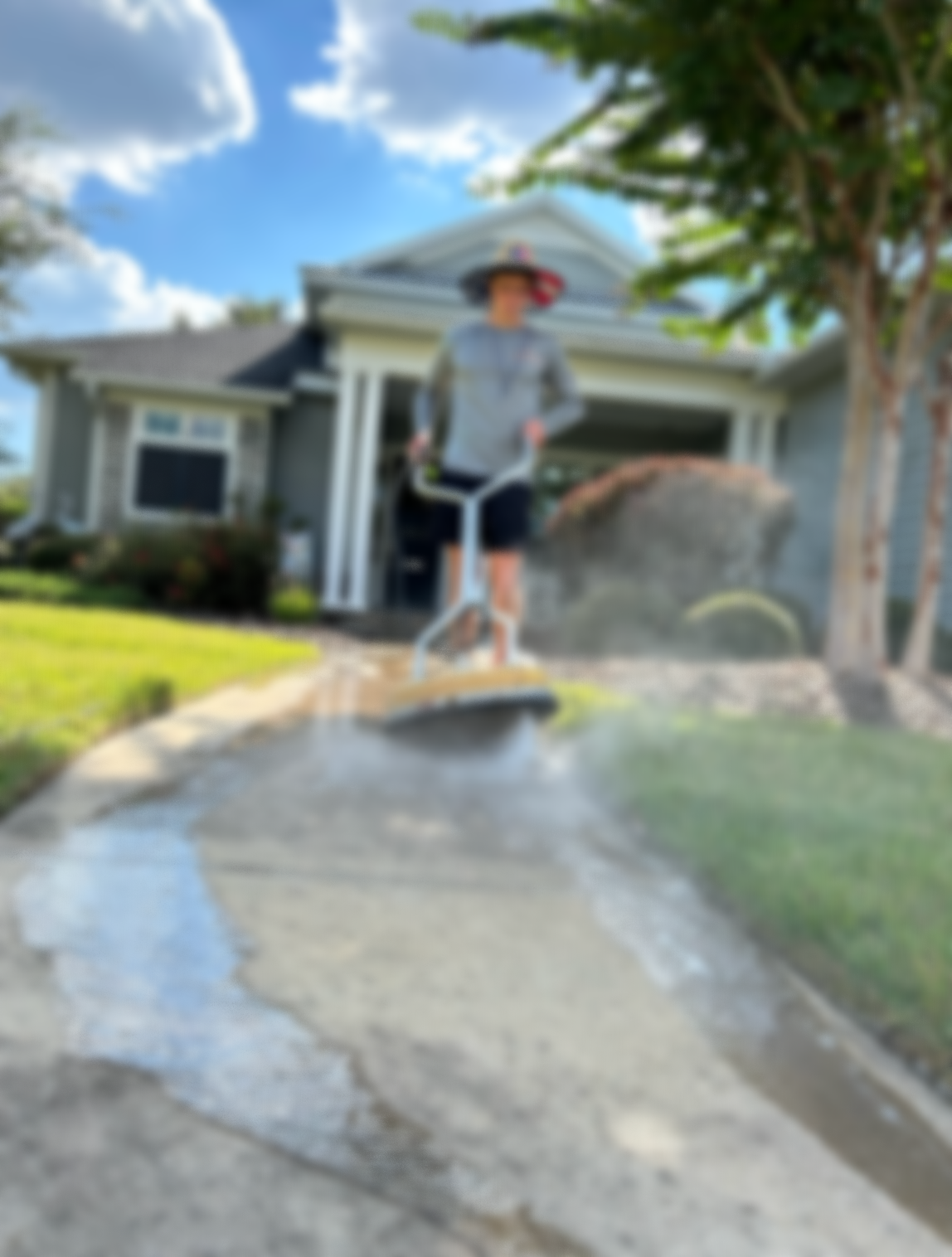 Person pressure washing a concrete sidewalk in front of a house on a sunny day.