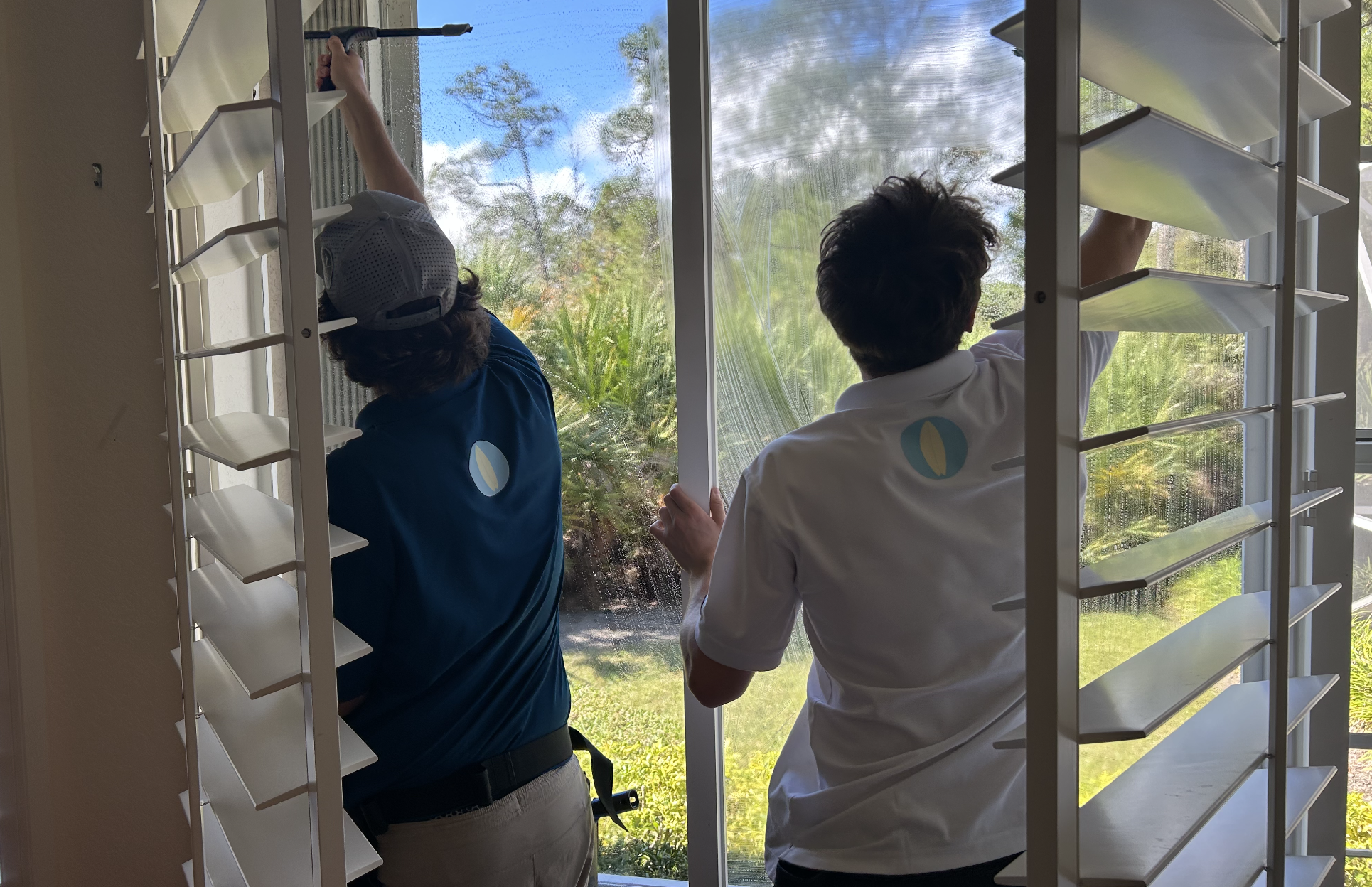 Two workers cleaning a large window from the inside, with a garden visible outside.