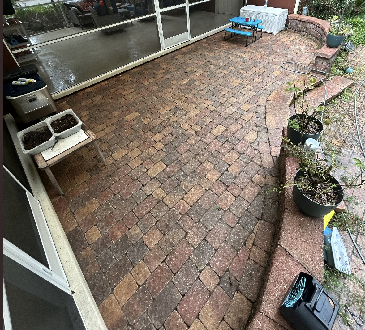 A brick patio with potted plants and a small blue table with chairs on the right side, wet from rain, with a screened porch and outdoor storage furniture in the background.