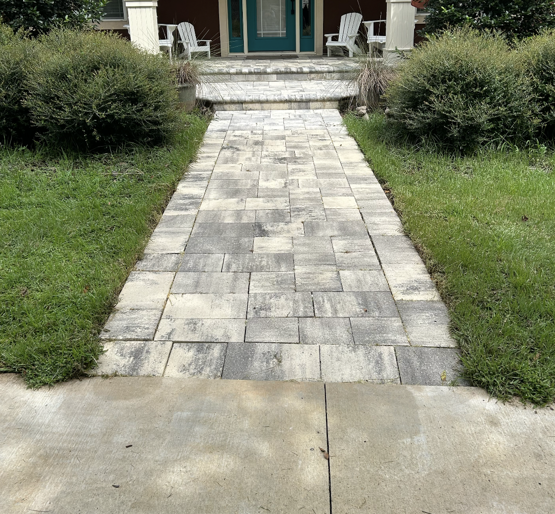 A stone pathway leading to the front porch of a house, flanked by green bushes and grass on both sides.