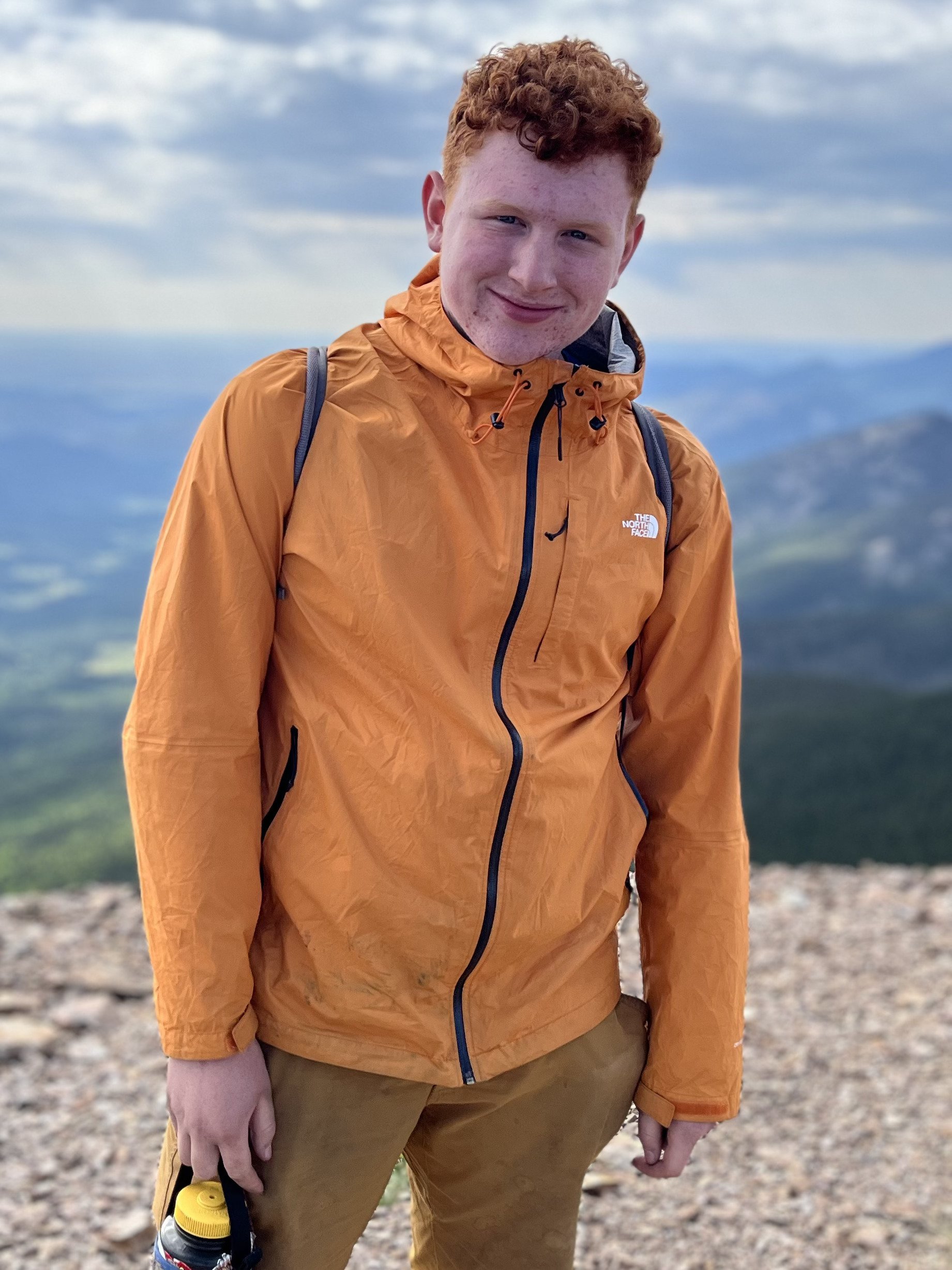Young man with curly red hair wearing an orange North Face jacket, carrying a small backpack, standing on a mountain with a scenic view of valleys and distant mountains in the background.