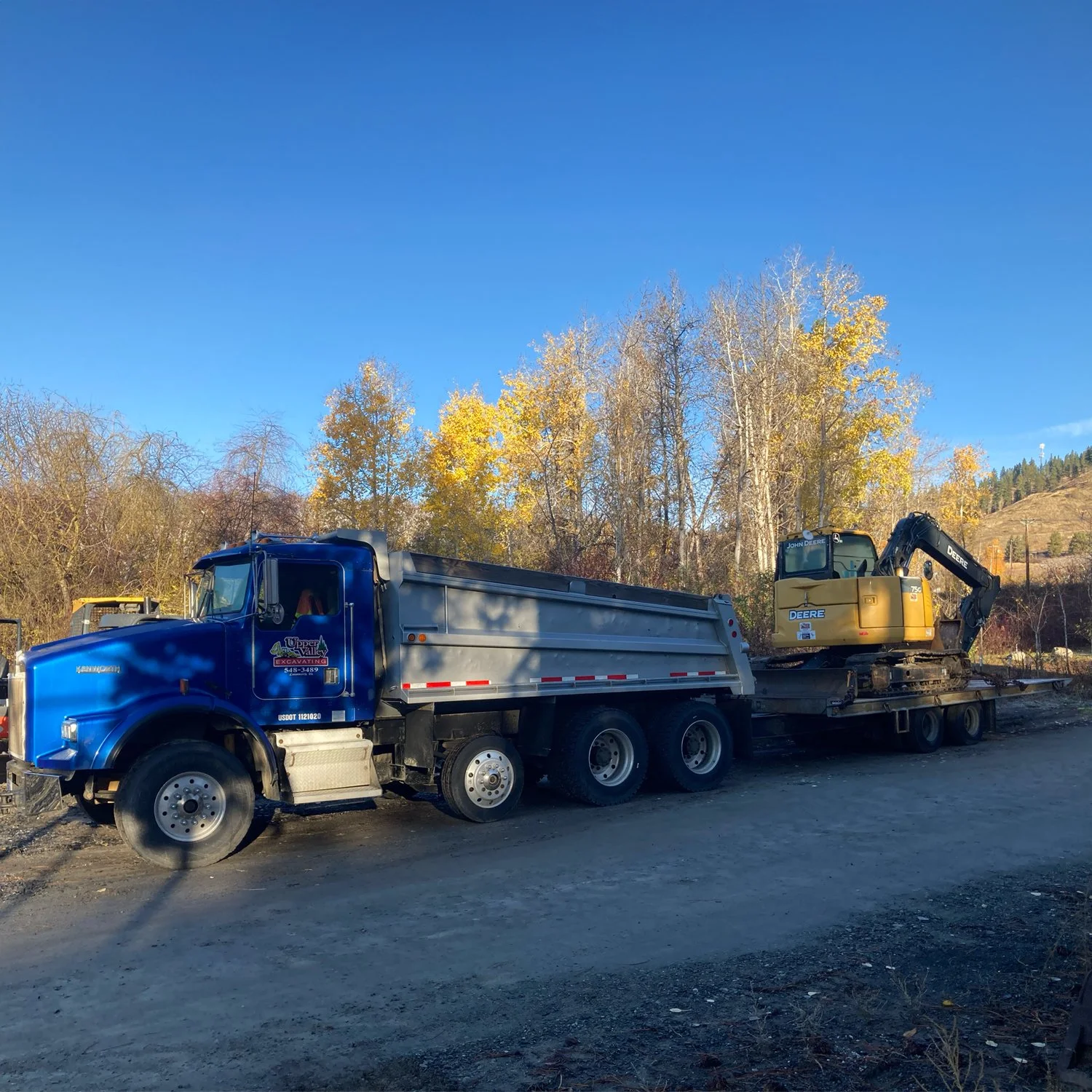 A large blue dump truck with a yellow and black excavator on its flatbed, parked outdoors on a dirt surface with trees and hills in the background.