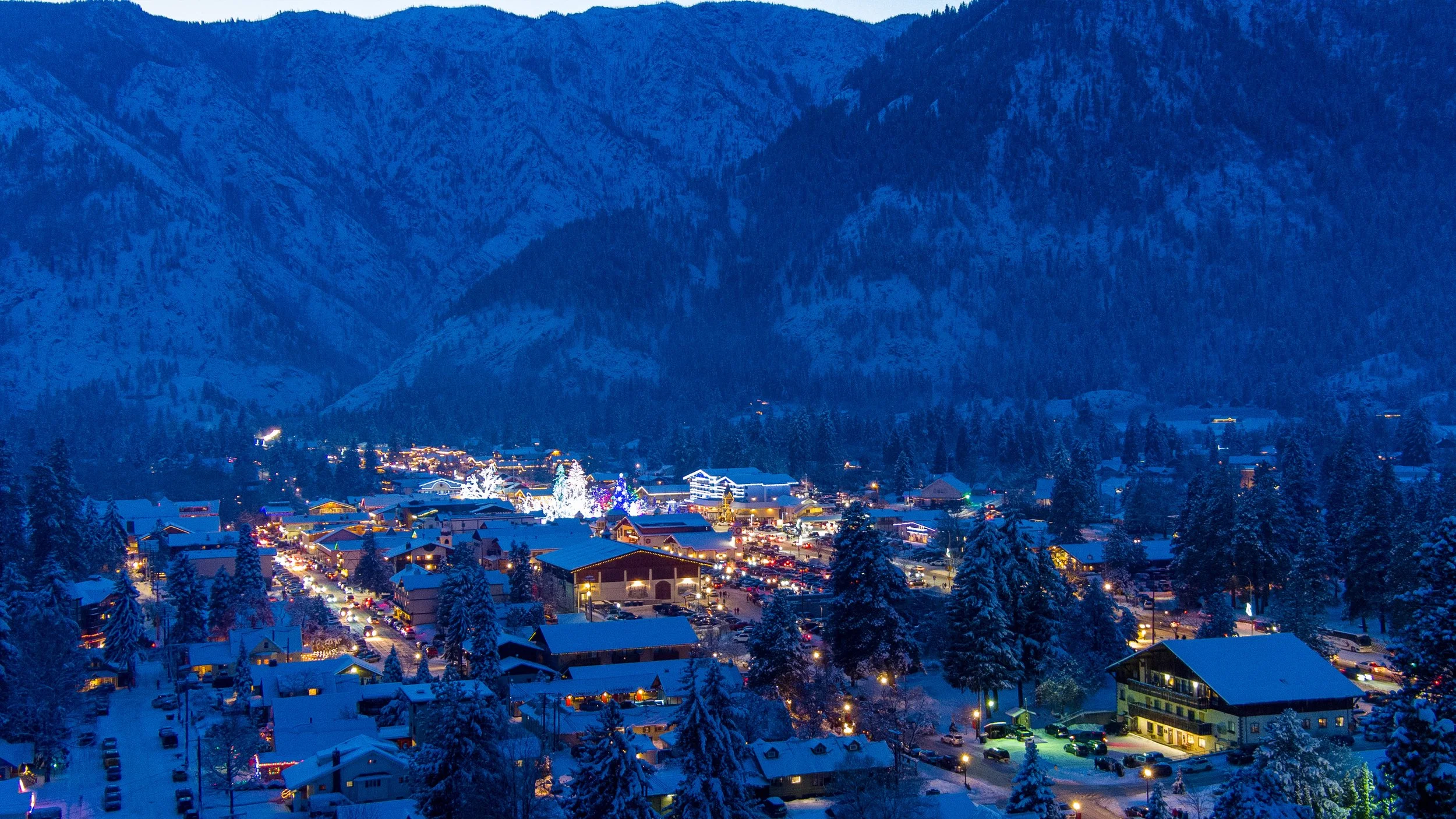 A snowy mountain town at dusk with festive holiday lights and decorated trees among houses and buildings, surrounded by forested mountains.