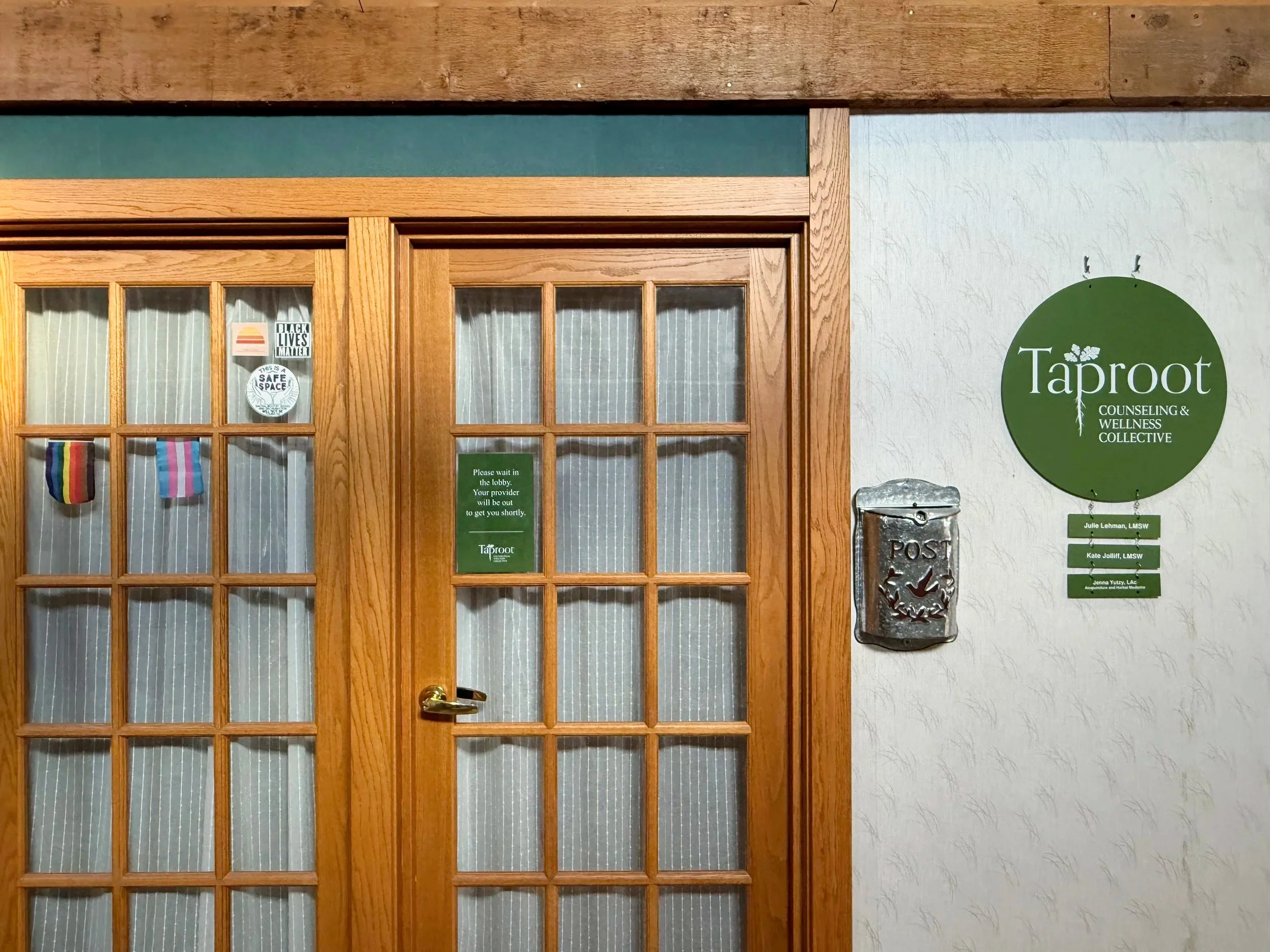 The main entrance to Taproot Acupuncture & Herbal Medicine in Newton, KS. The glass doors feature inclusive Pride flags, and the wall display shows the Taproot Counseling & Wellness Collective sign with individual practitioner names hanging below.