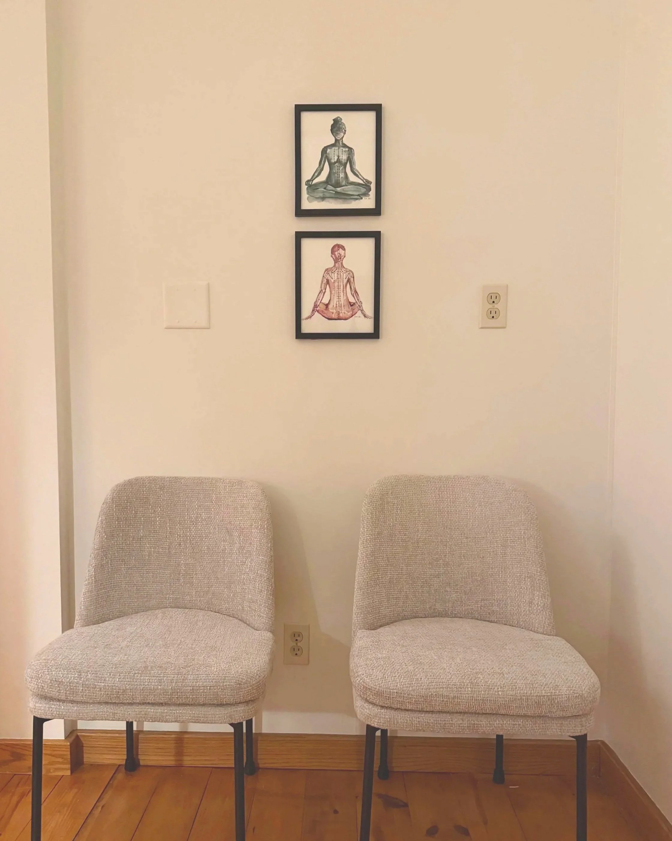 Two comfortable grey chairs in the patient intake area, positioned beneath framed watercolor artwork of acupuncture channels on the human body.