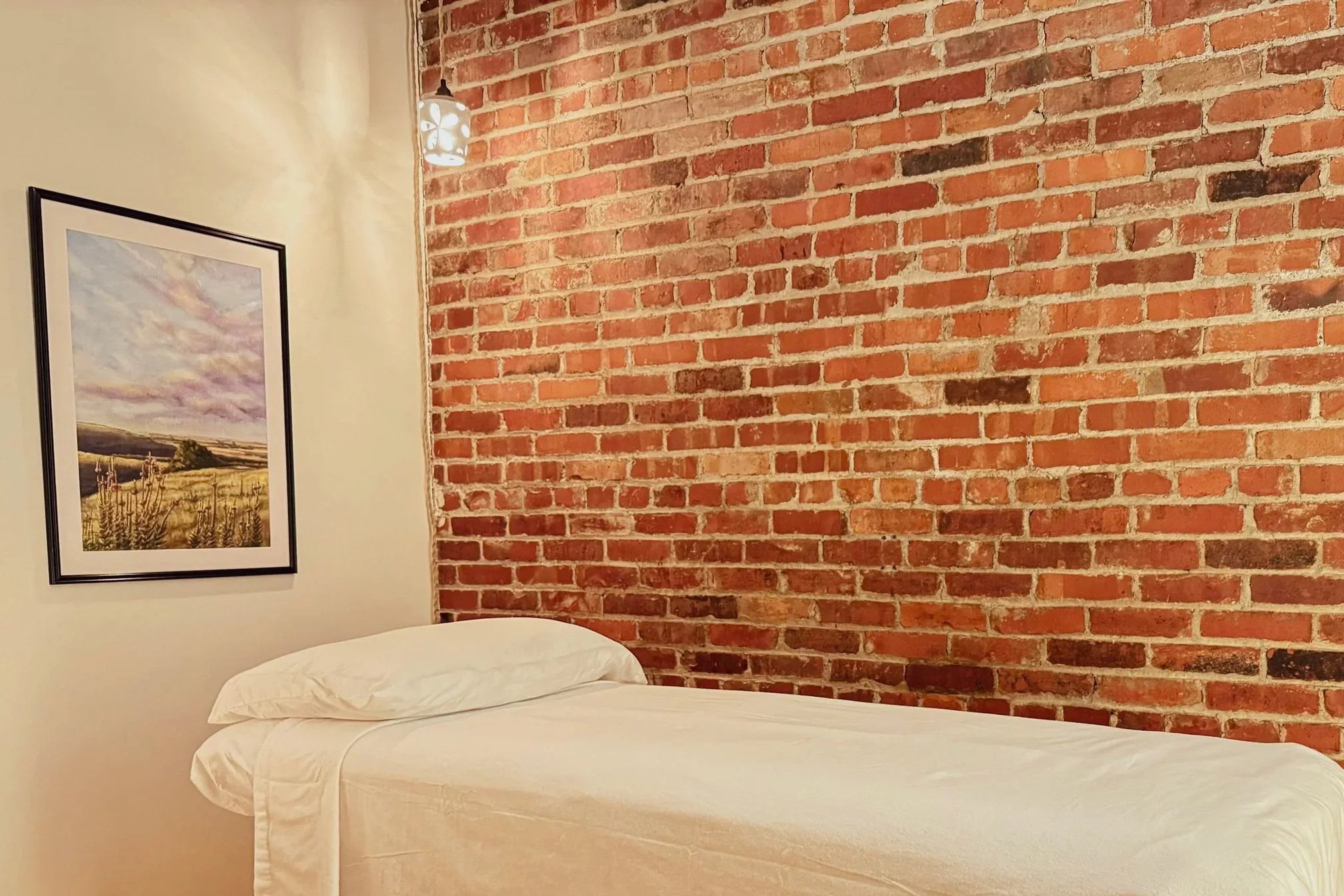 A comfortable acupuncture treatment table with crisp white linens set against a historic red brick wall and soft ambient lighting at our Newton, KS clinic.