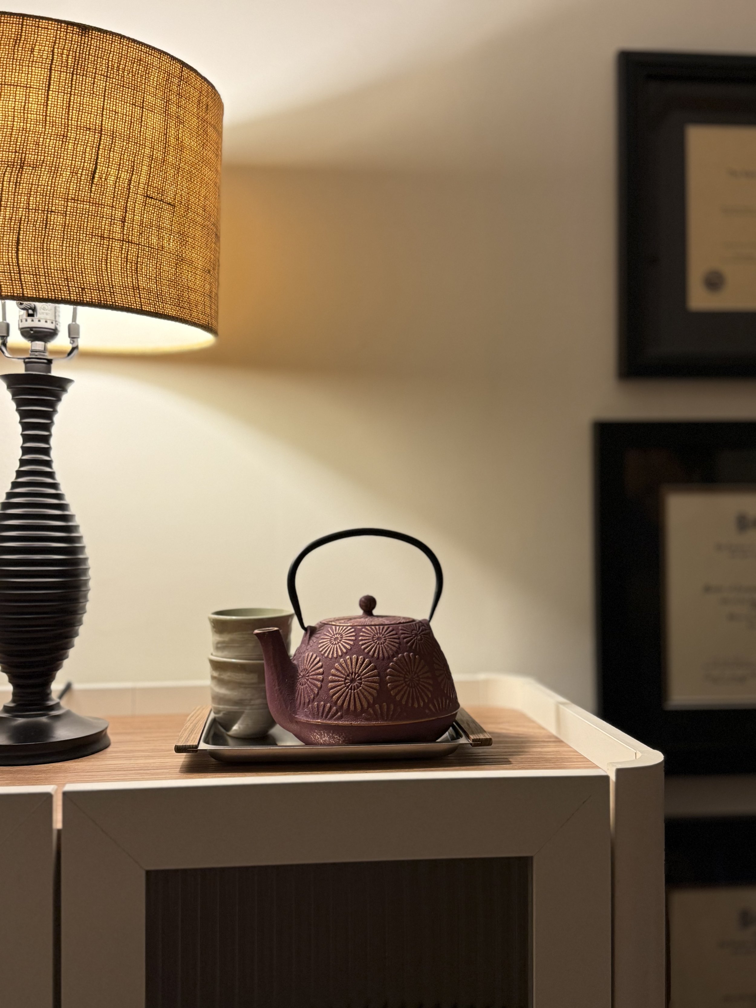A warm, inviting scene at Taproot Acupuncture & Herbal Medicine featuring a cast iron teapot on a wooden cabinet and soft lighting, with a glimpse of Jenna Yutzy's framed medical diplomas and credentials in the background.