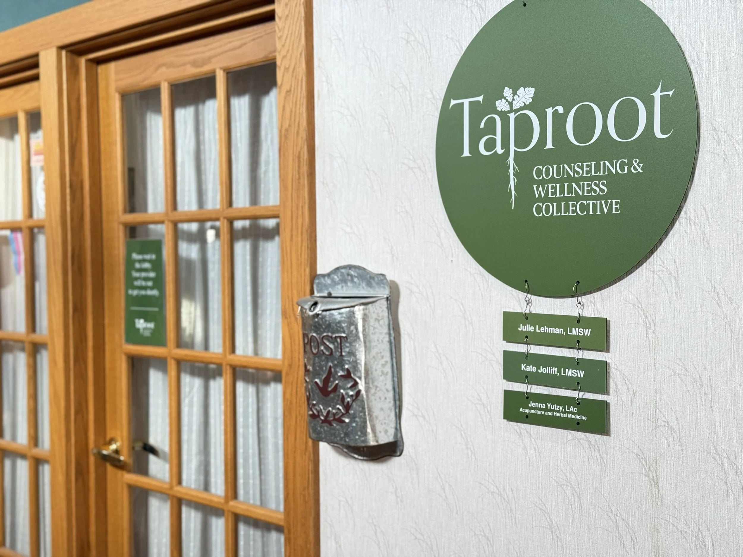 A close-up view of the entrance to the Taproot Counseling & Wellness Collective suite, featuring the green circular logo on the wall with individual practitioner names hanging below it. The entrance includes a set of wooden, glass-paneled doors.