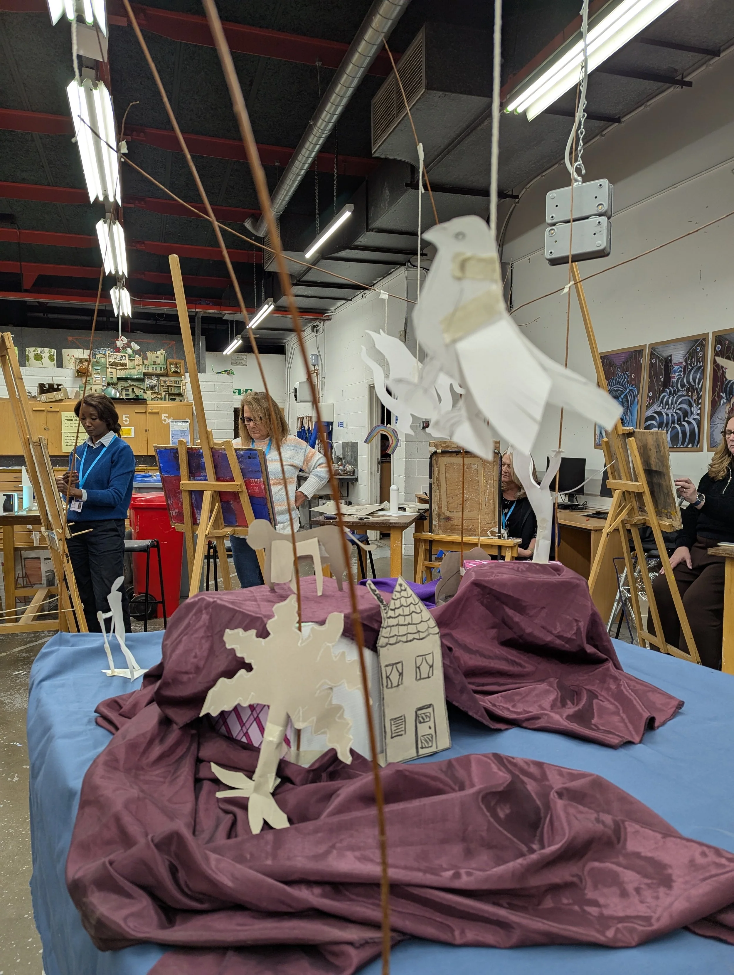Scene of a puppetry or puppet-making workshop with crafted paper puppets of a snowman, dragon, house, palm tree, and unicorn hanging from strings over a table draped with purple fabric. In the background, three women are working at easels or tables i