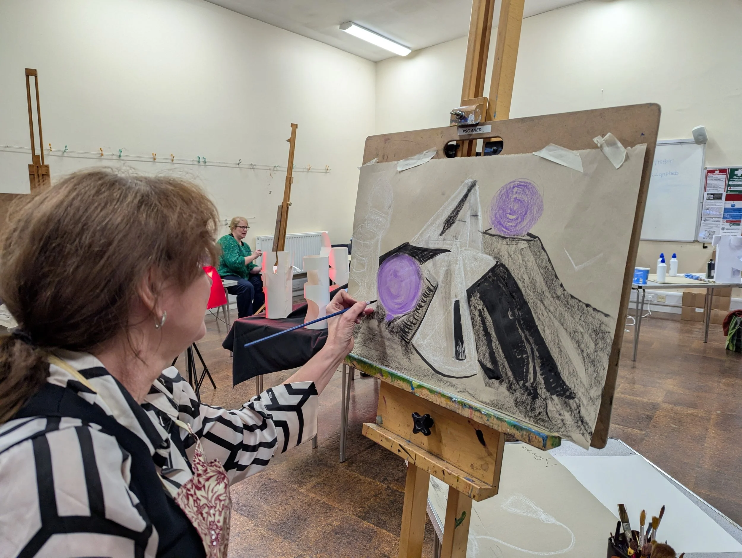 Student painting abstract art on a canvas in an art class or workshop, with other participants and art supplies visible in the background.