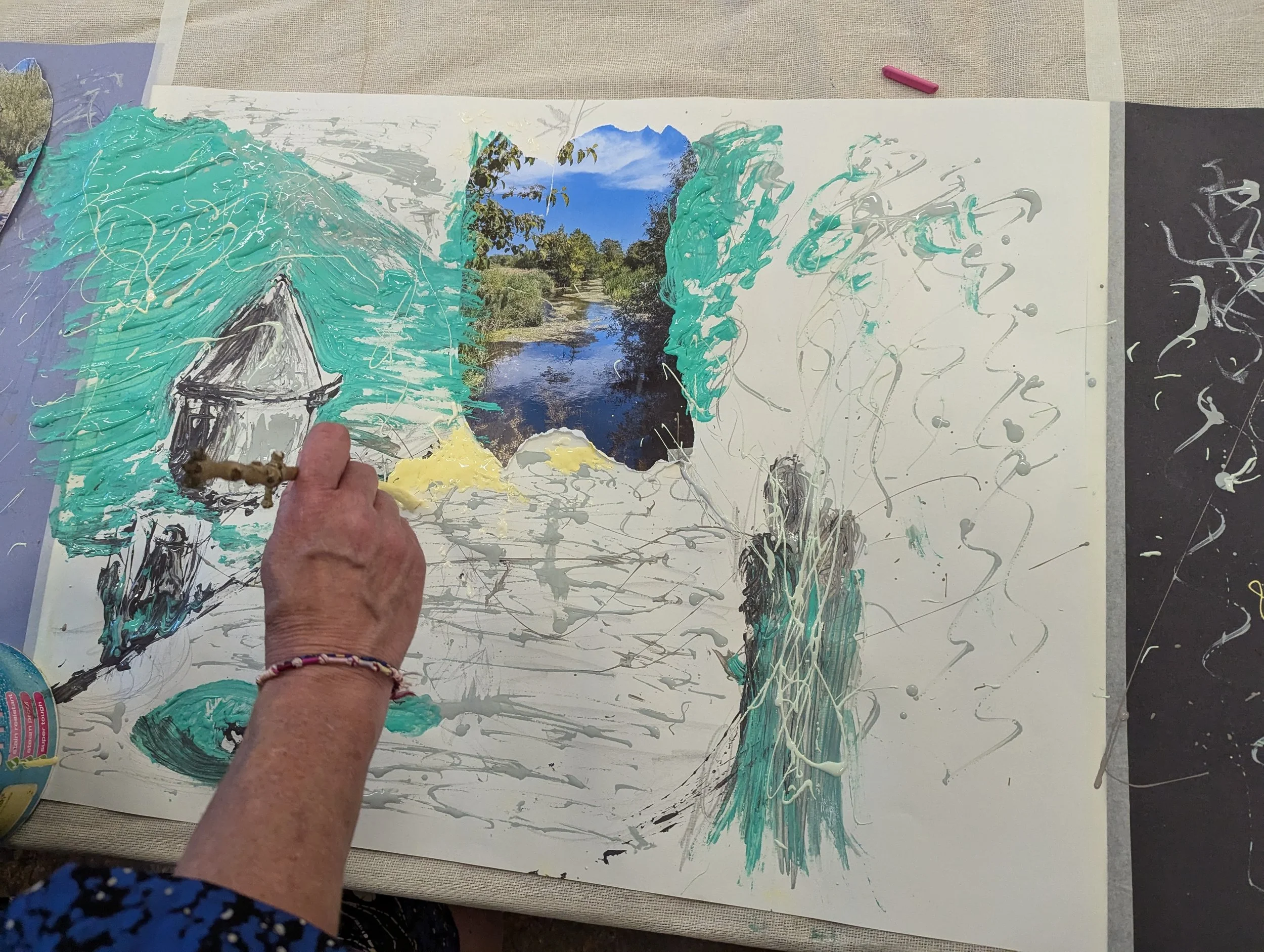 Student working on a mixed-media landscape painting that includes a photograph of a river and hills, with painted trees, a small house, and abstract splashes of paint.