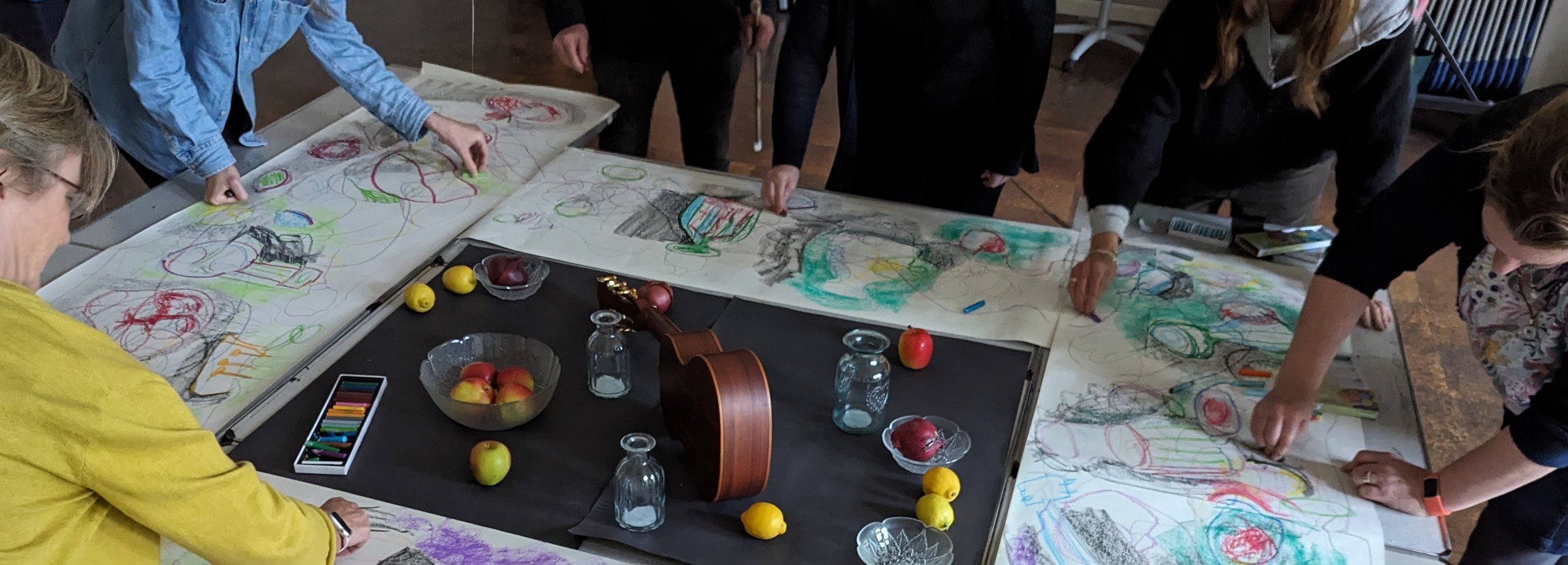 Students gathered around a large table, drawing and coloring on long sheets of paper with various art supplies, fruits, and glassware on the table.