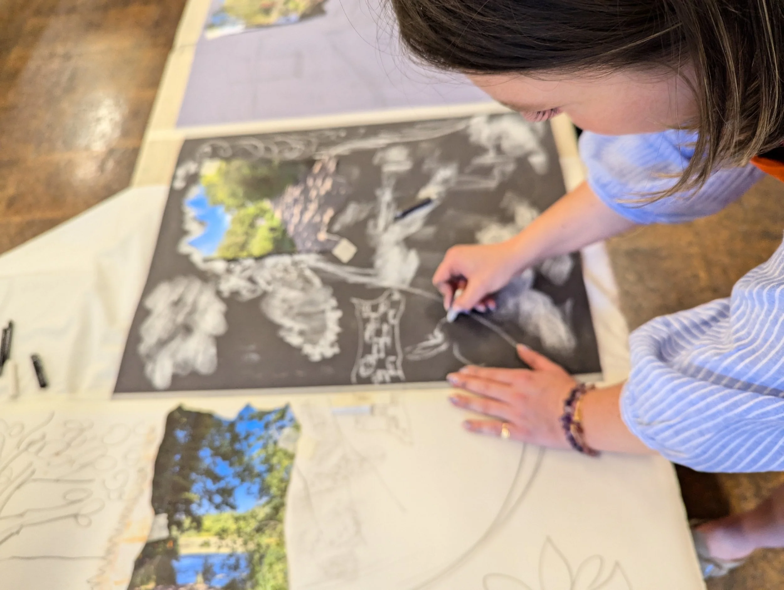 Student drawing a landscape scene on black poster with white chalk, with another landscape image underneath on paper.