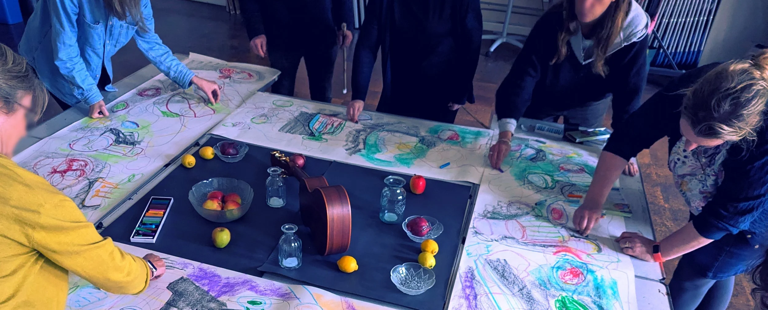 Group of people drawing and coloring on large sheets of paper spread across a table, with apples and glass jars on the table.