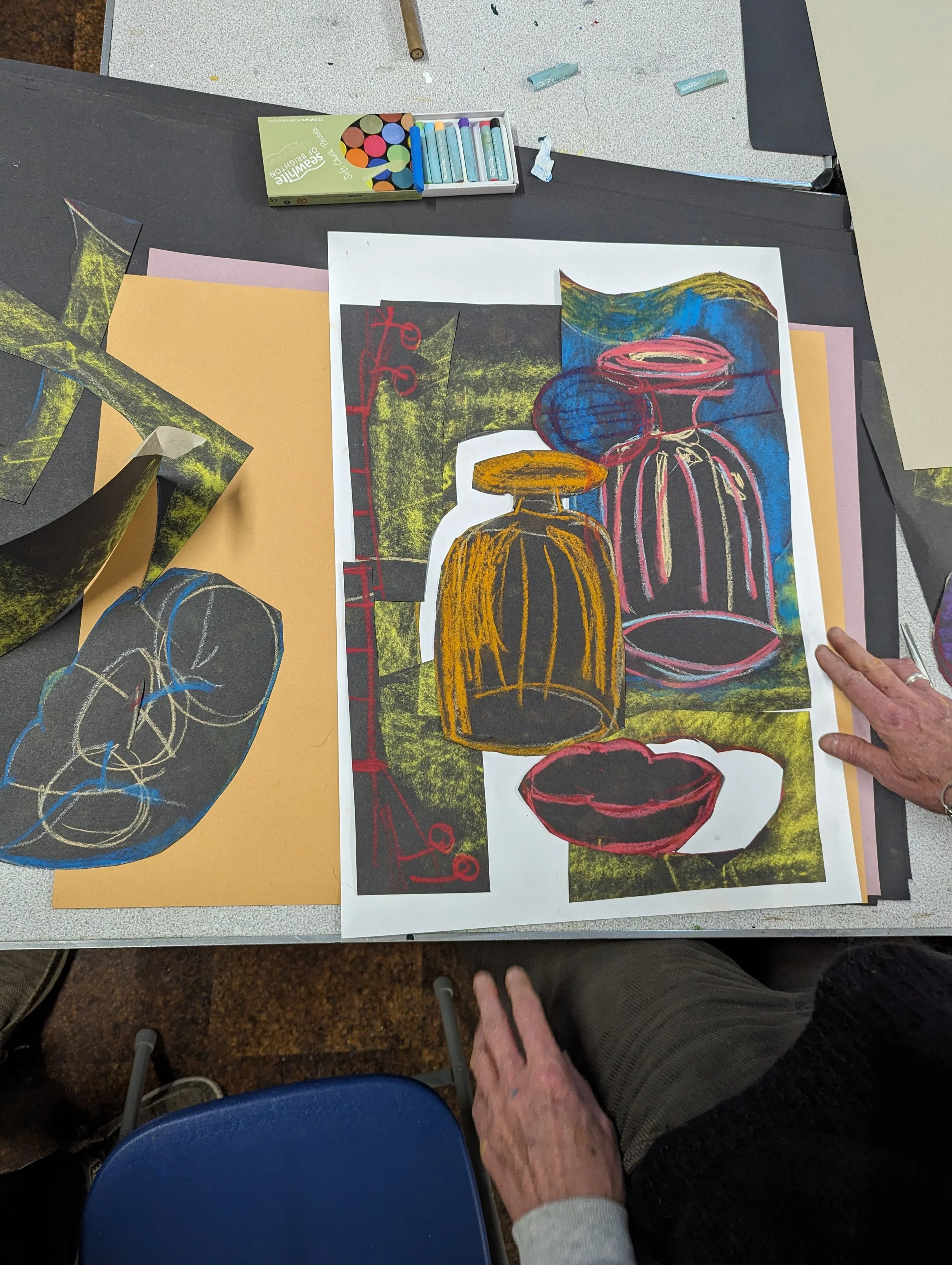 Colorful artwork of bottles and bowls on a table, with cutouts and sketches, an individual working on the art piece.