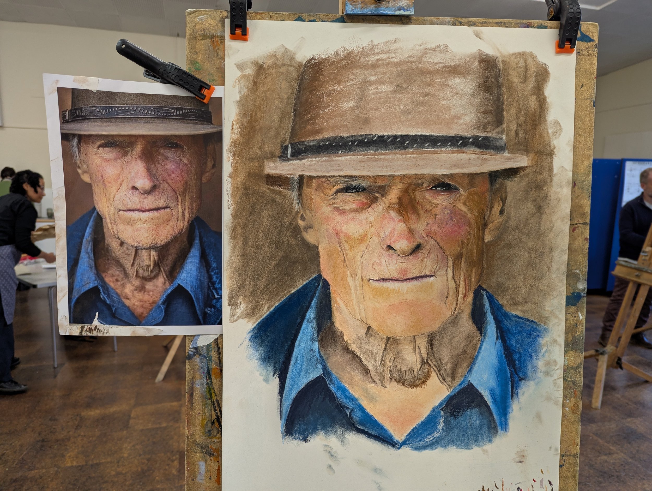 An unfinished pastel drawing of an elderly man…..Klint Eastwood!!…. in a blue shirt and gray hat, with the reference photo of the man in the background on the left.