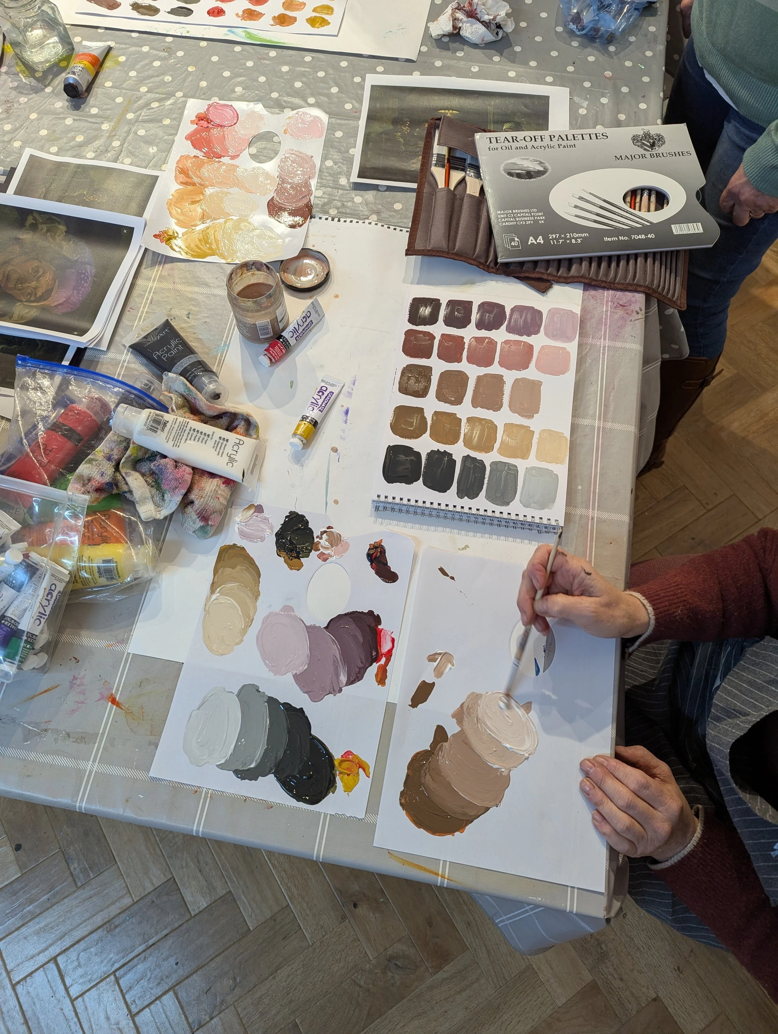 A table with various paint palettes, brushes, and color swatches used for painting. A person is mixing paint on a palette, and there are sample color sheets and painting supplies scattered across the table.