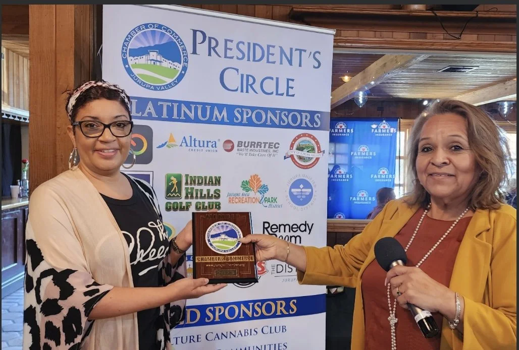 Two women standing in front of a banner at a business event. The woman on the left is smiling, holding a plaque, and wearing glasses, a black shirt with white writing, and a patterned shawl. The woman on the right is holding a microphone, wearing a y