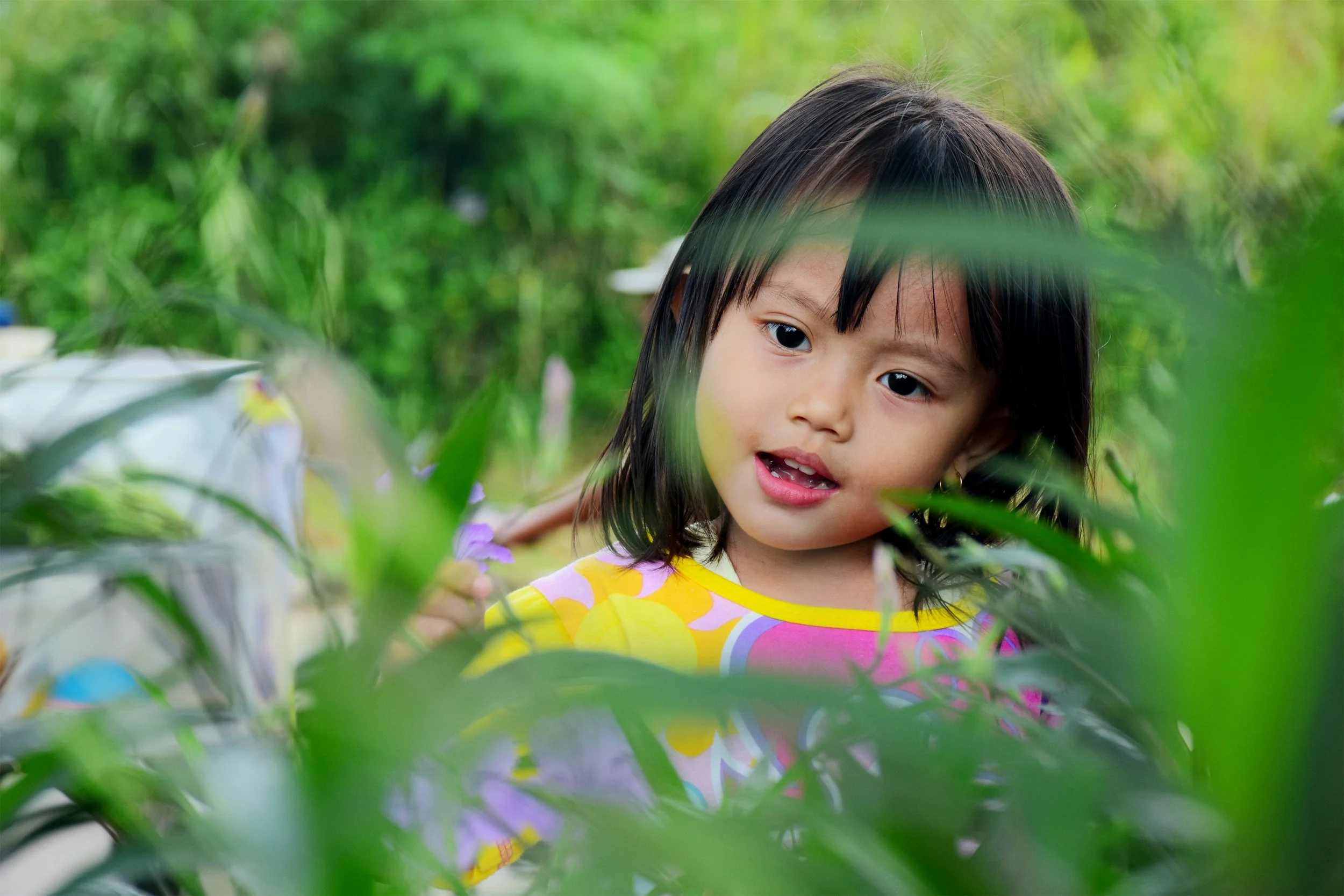 A young girl with dark hair, wearing a colorful dress, is looking through green plants outdoors in a lush, green environment.