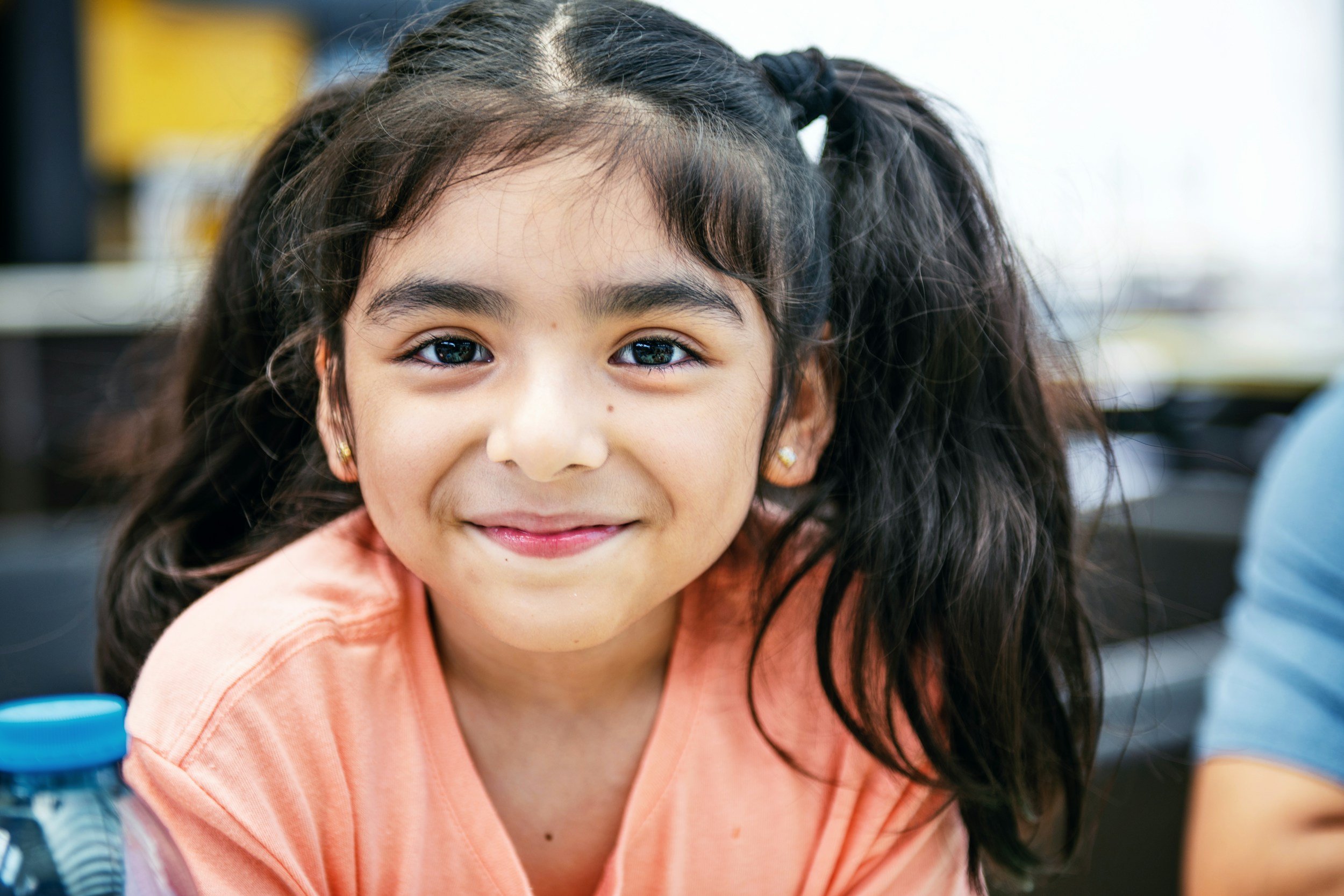 Close-up of smiling young girl with dark curly hair and earrings, wearing an orange shirt, indoors with blurred background.