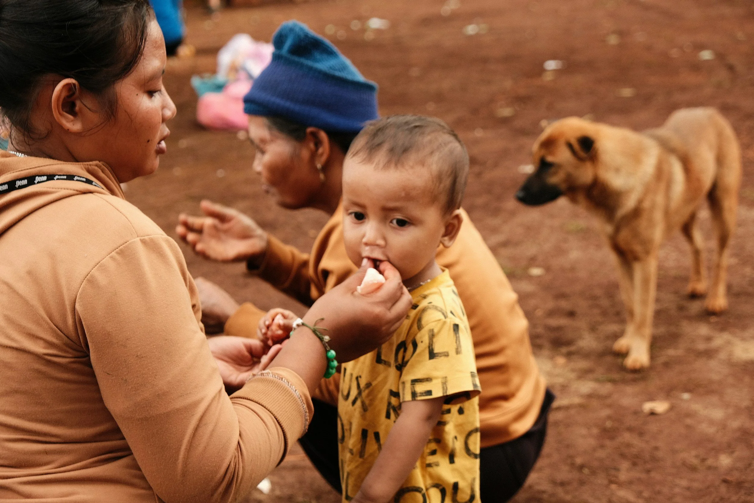 A woman, child, and dog on a dirt ground, with the woman handing something to the child, who is looking at her. A dog stands nearby, and there are blurred objects in the background.