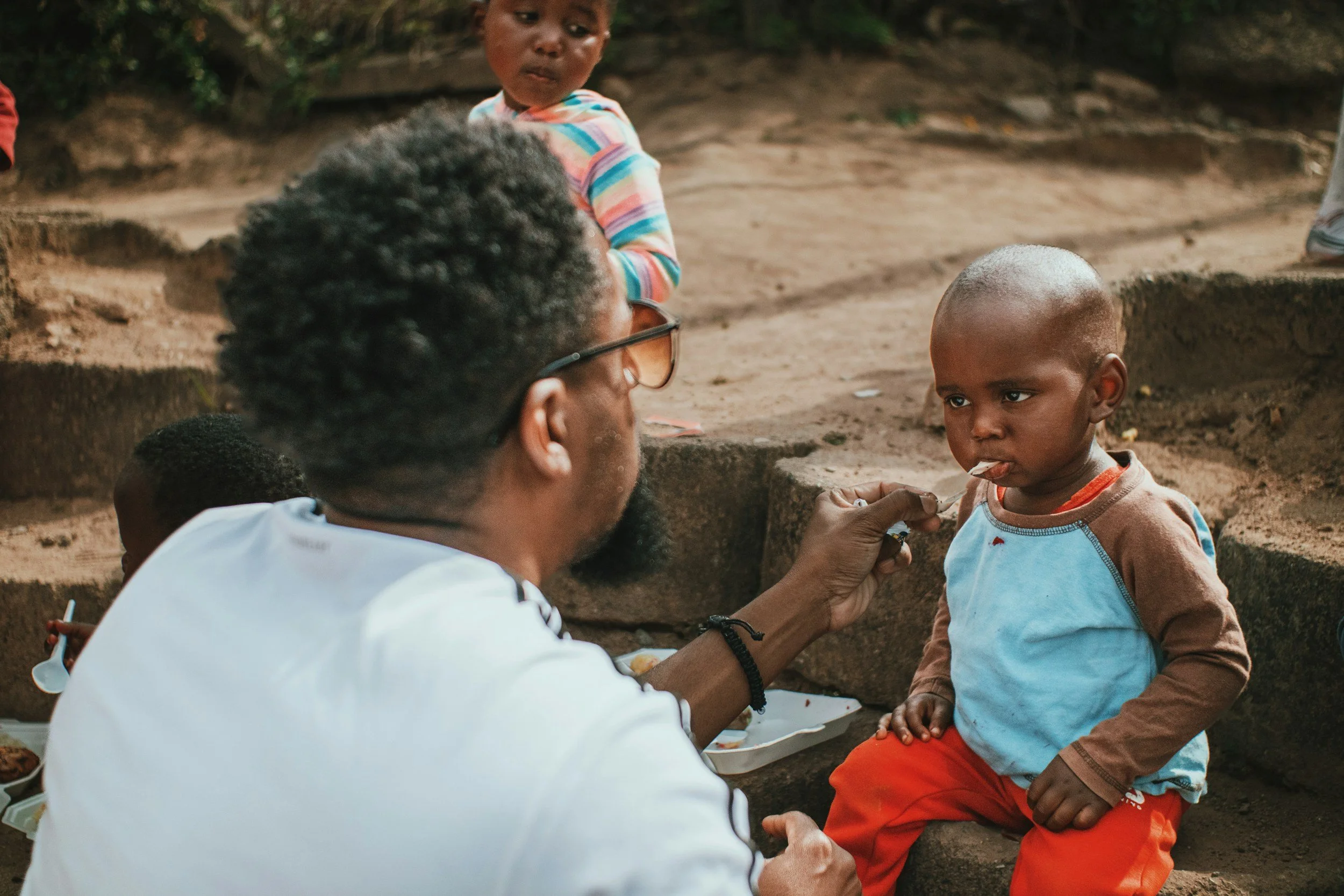 A young boy sitting outdoors with a serious expression, wearing an orange and blue shirt and orange pants, as an adult man with glasses and a beard examines or holds his hand. Another child with a striped shirt stands nearby, and parts of other children are visible in the background.
