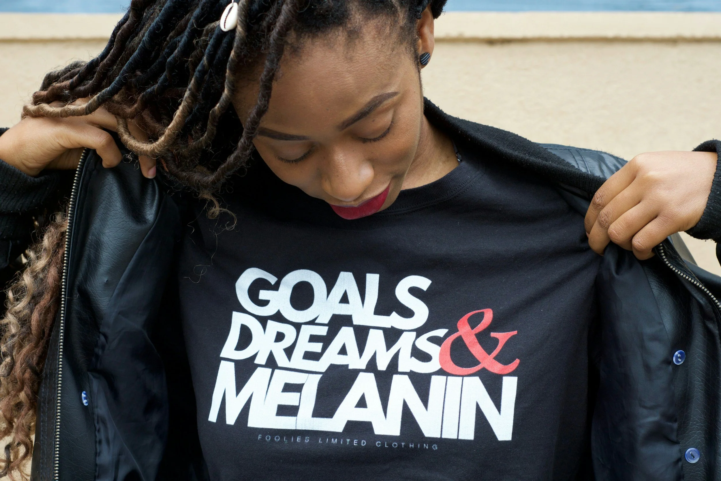 A close-up of a woman with long, curly dreadlocks, wearing a black leather jacket and a black t-shirt with bold white and red text. She is looking down while adjusting her jacket.