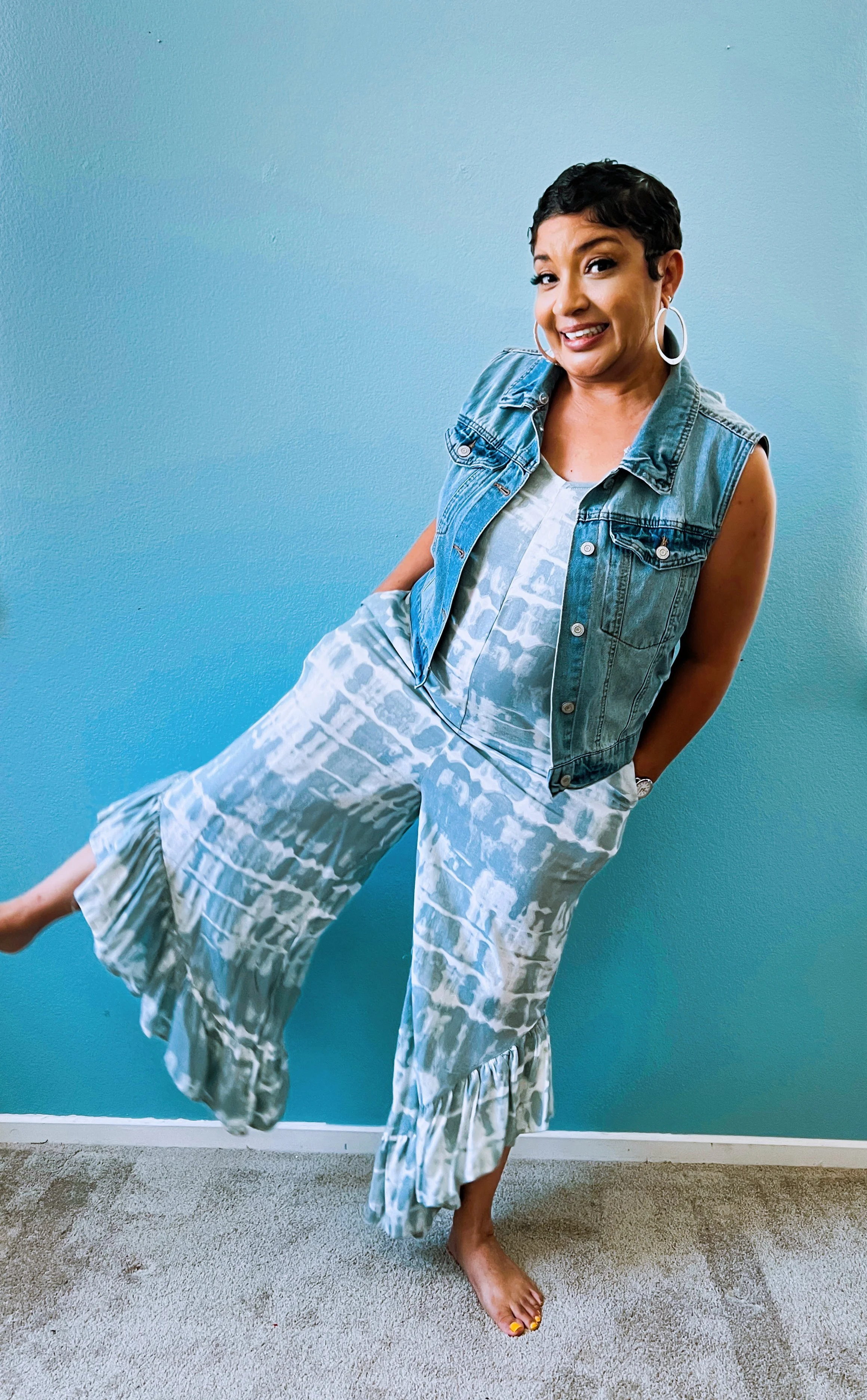 Woman with short dark curly hair wearing large hoop earrings, denim vest, patterned dress, and patterned pants, standing against a blue wall with one leg lifted and smiling.