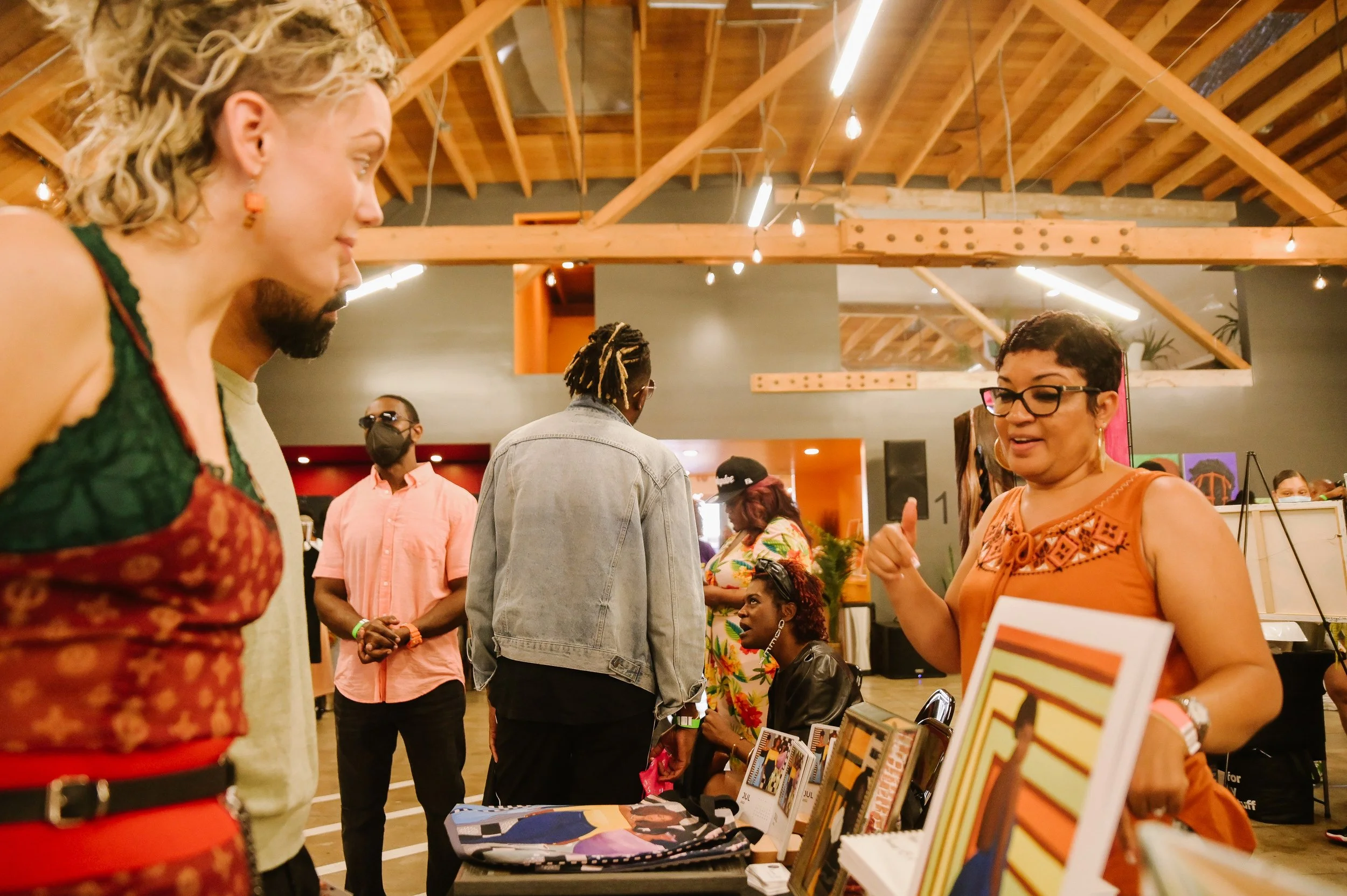 People gathered indoors, engaging in conversation at a booth with books and artwork, with wooden beams and warm lighting in the background.