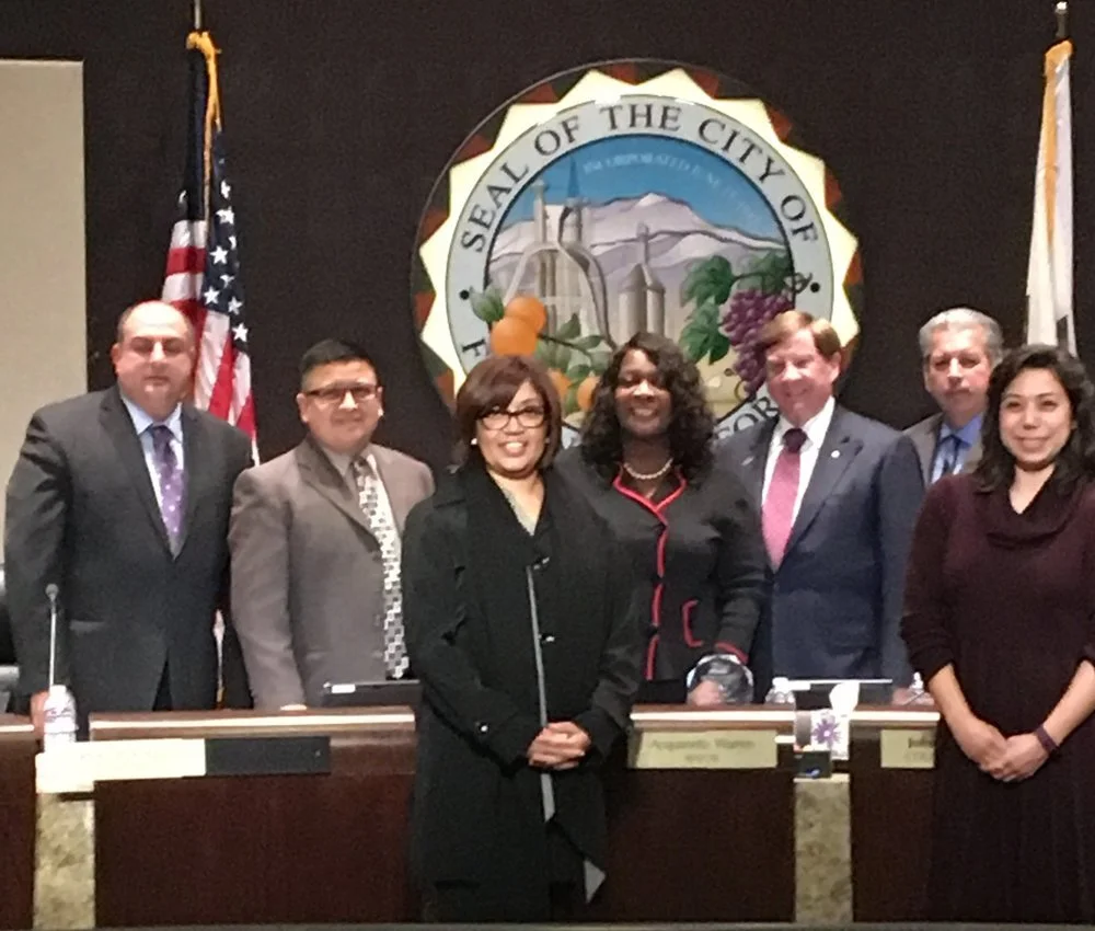 Group of six people standing in front of a city seal and flags in a formal setting.