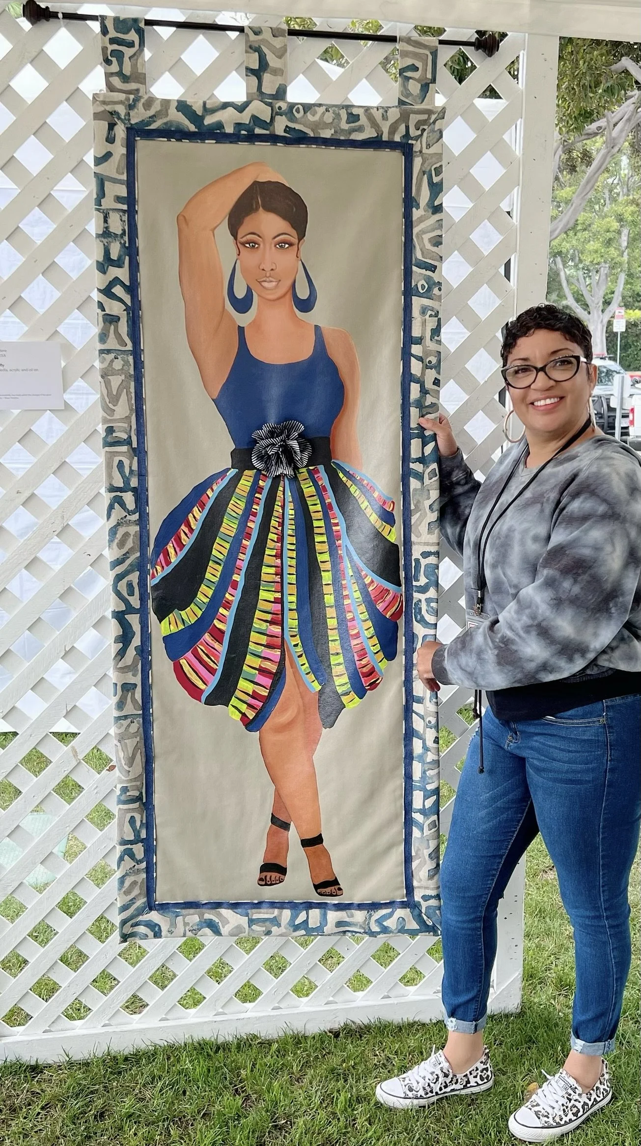 A woman with short dark hair and glasses stands next to a colorful painting of a woman in a blue top and vibrant skirt, posing with one arm above her head against a white lattice background.