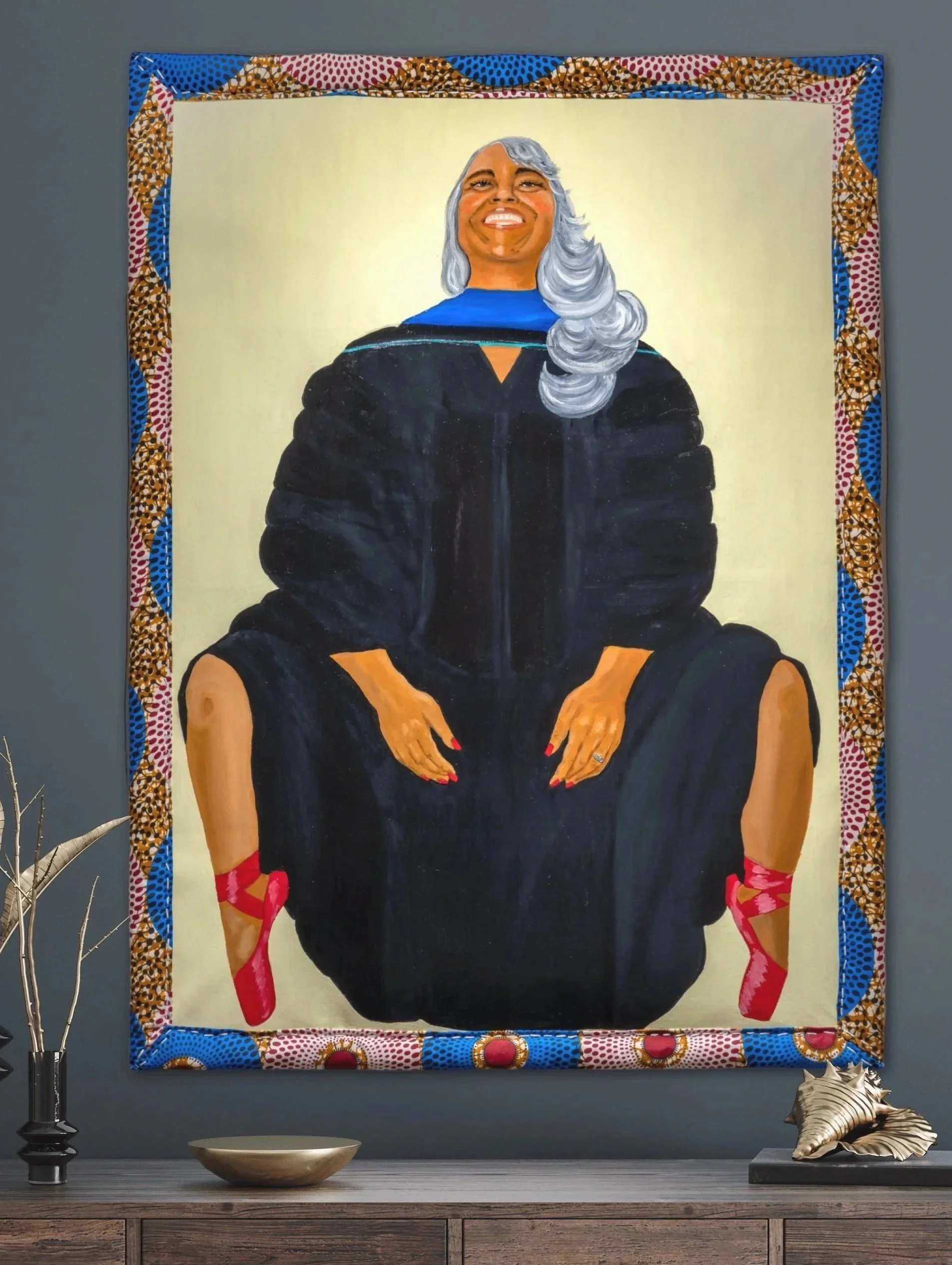 A colorful painting of a woman with silver hair, wearing a graduation gown, seated with her hands in her lap, framed with bright patterned fabric, displayed on a wall in a room with decorative objects on a table below.