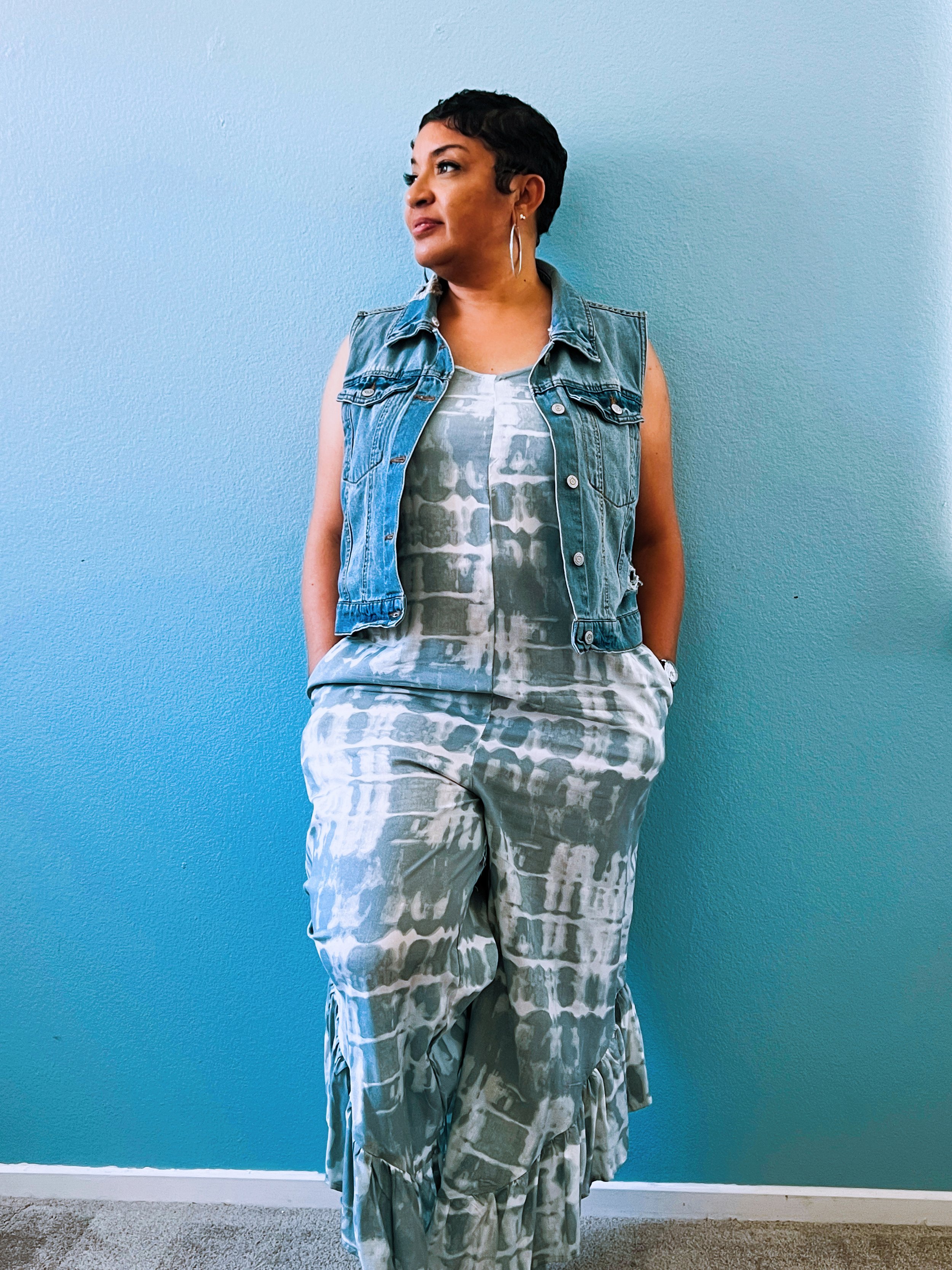 A woman with short dark hair, wearing large hoop earrings, a sleeveless denim vest, a white and grey patterned dress, and matching wide-leg pants, standing against a blue wall.