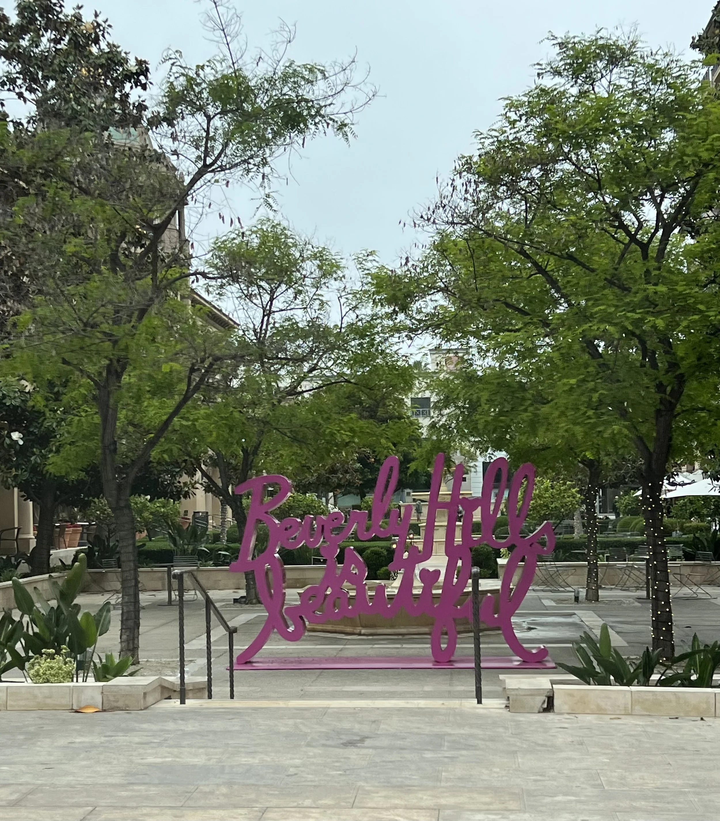 Decorative pink sign with cursive writing in front of a park scene, trees, buildings, and cloudy sky in the background.