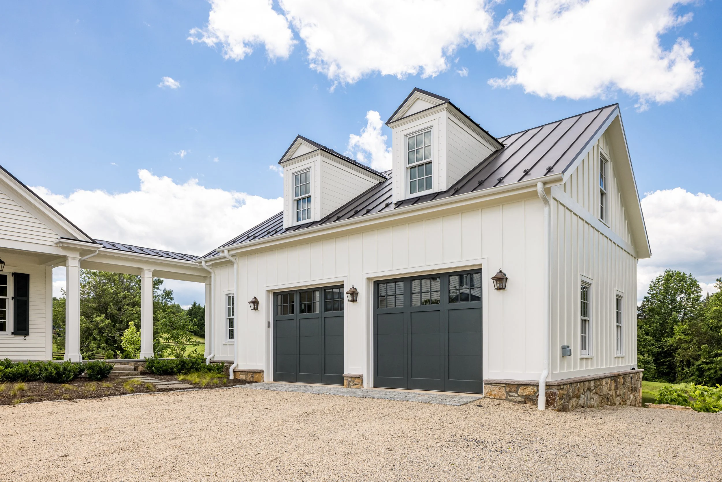 A white house with black window frames and a metal roof, featuring a two-car garage with dark gray doors, surrounded by a gravel driveway and greenery, under a partly cloudy sky.